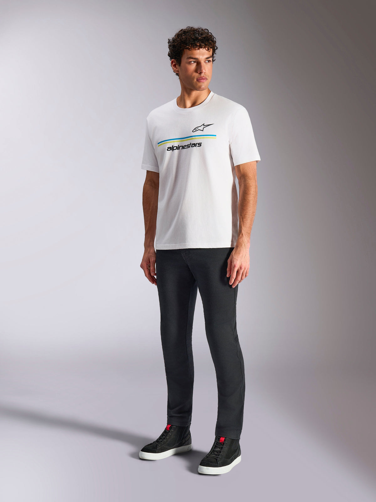 Alpinestars Lanes CSF Tee, White Short Sleeve T-Shirt, worn by model with black slim-fit pants and black high-top sneakers, featuring blue and yellow horizontal stripes with Alpinestars branding on the chest, casual motorsport-inspired streetwear