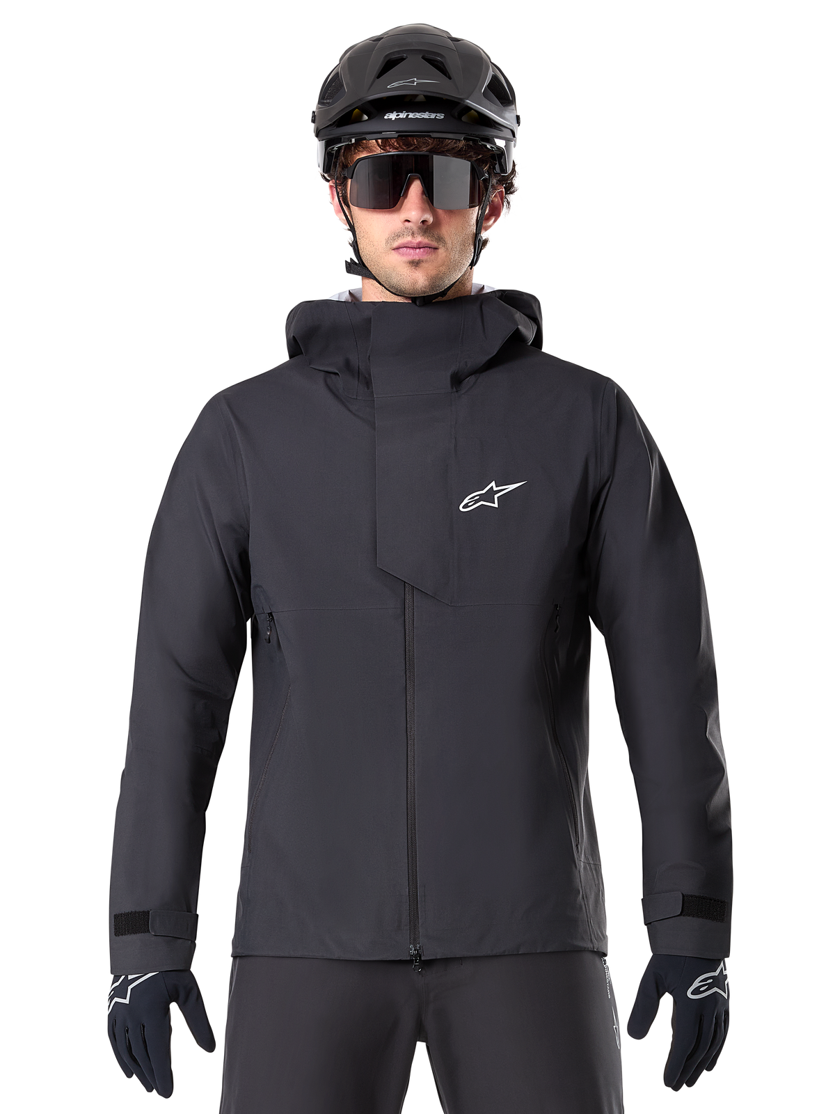 Alpinestars A-Dura Elite Wasserdichte Jacke, Schwarz, Mountainbike-Jacke, Design mit hohem Kragen und Kapuze, Frontreißverschluss mit schützender Brustklappe und weißem Logo, getragen mit schwarzem Helm und Fahrradhandschuhen, technischer Wetterschutz für Trail-Fahrten