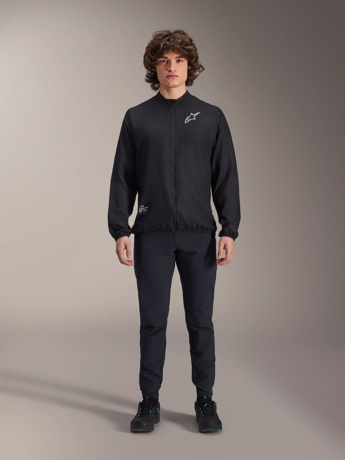 Model trägt die Alpinestars A-Aria Windbreaker-Jacke in Schwarz, mit durchgehendem Front-Reißverschluss, Stehkragen und weißem Astars-Logo auf der Brust, kombiniert mit schwarzer Funktionshose und schwarzen Schuhen, Performance-Radbekleidung für Mountainbiking und Outdoor-Aktivitäten.