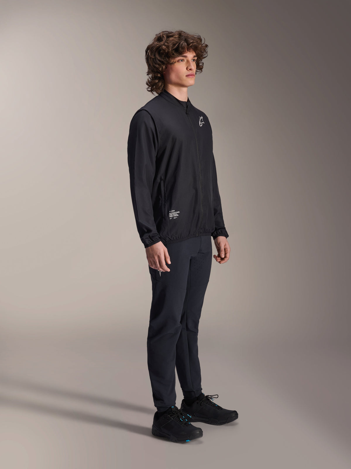 Alpinestars A-Aria Windbreaker Jacke, Schwarz, leichte windabweisende Radjacke, getragen von einem Model mit lockigem Haar, weißes Astars-Logo auf der Brust und Branding unten rechts, kombiniert mit schwarzer Hose und Schuhen, Performance-Passform für Mountainbiking und Gravel-Riding