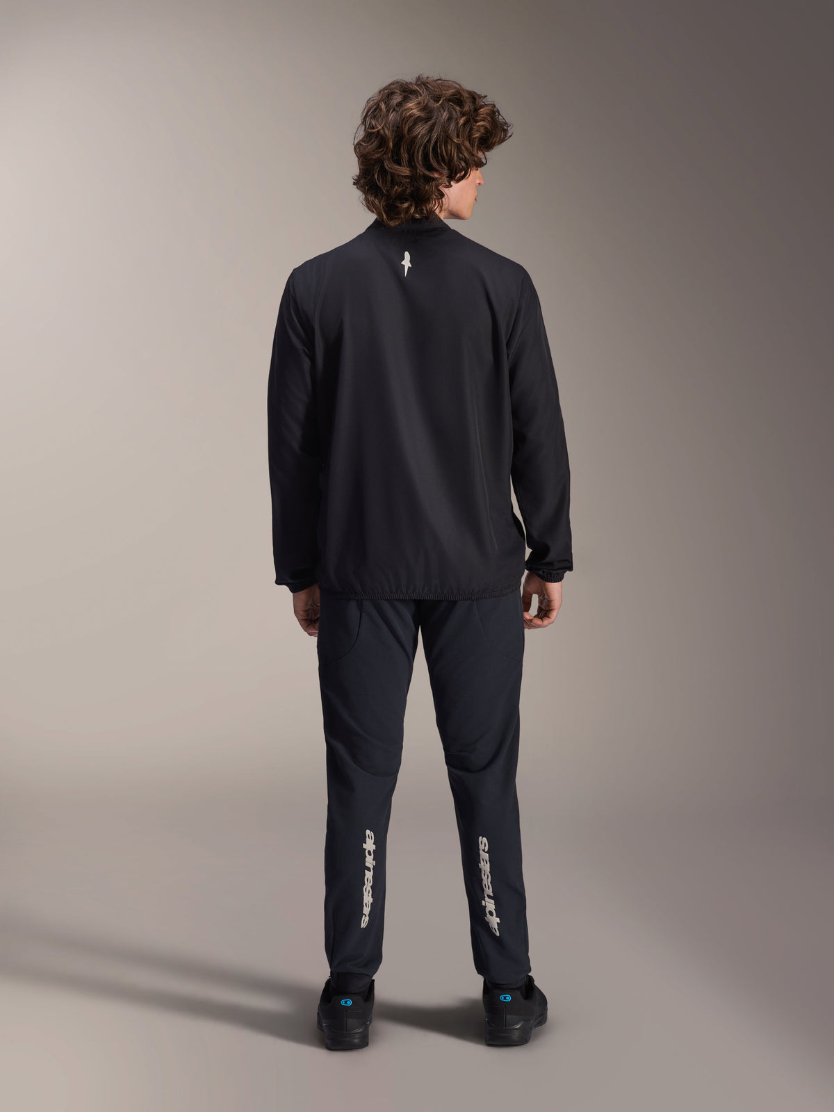 Alpinestars A-Aria Windbreaker-Jacke, Schwarz, Rückansicht, leichte windabweisende Radsportjacke mit einem kleinen weißen Logo auf dem oberen Rücken, getragen mit passender schwarzer Hose mit vertikalem Alpinestars-Branding an den Waden, Performance-Mountainbike-Ausrüstung