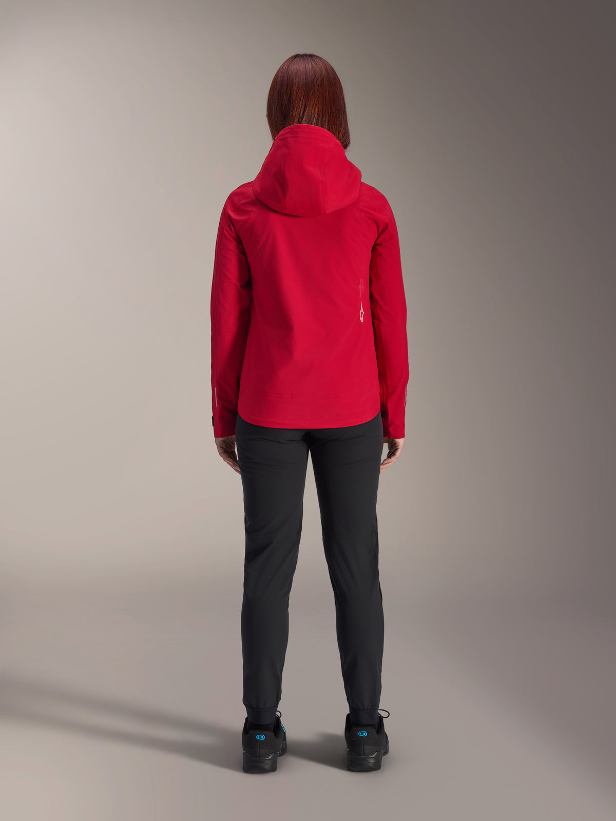 Back view of a woman wearing the Alpinestars Stella A-Dura Elite Waterproof Jacket in Red Fluo, styled with black pants and shoes, featuring a protective hood and dropped tail hem for mountain biking.