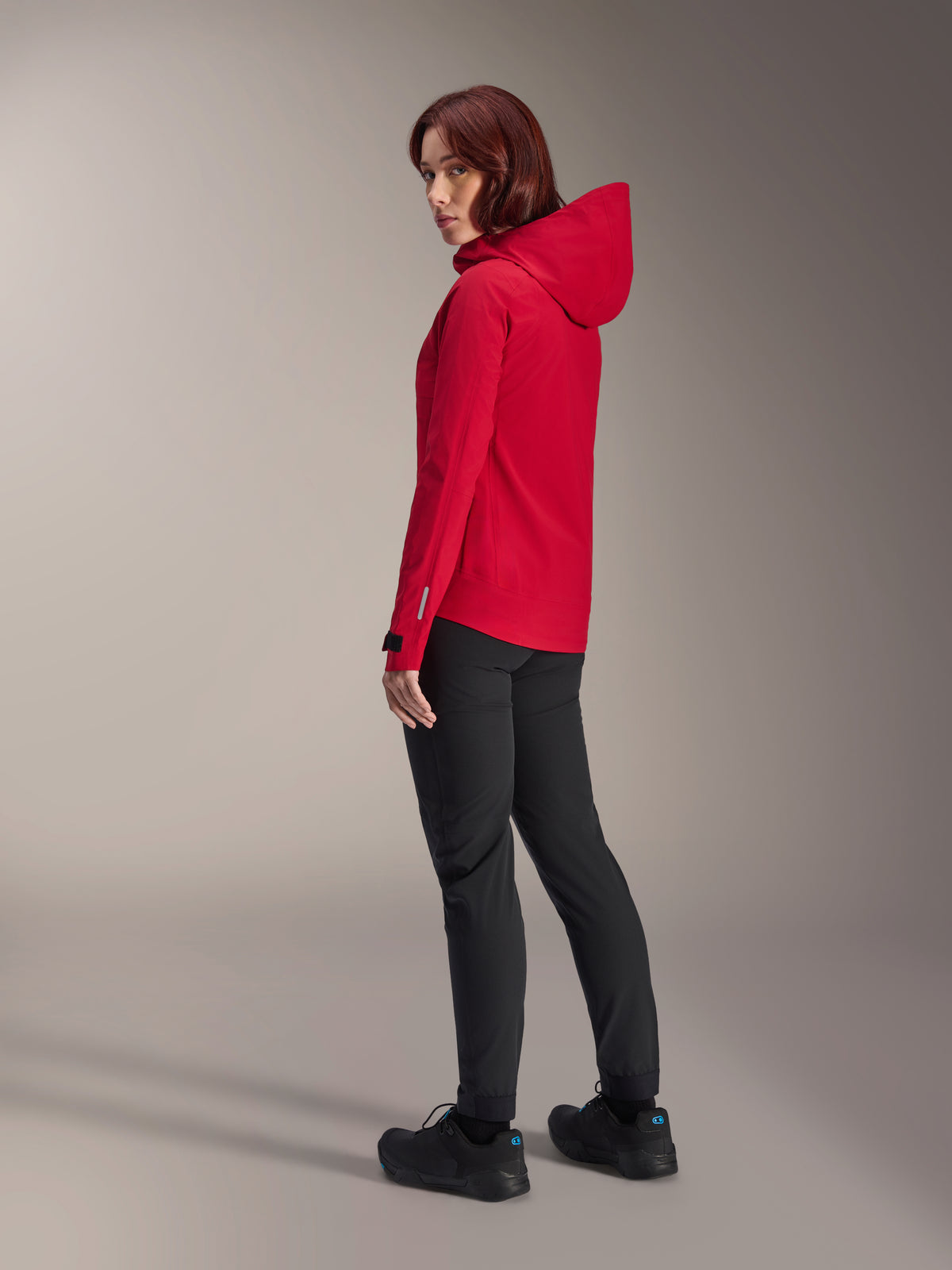 Woman wearing Alpinestars Stella A-Dura Elite Waterproof Jacket in Red Fluo, paired with black technical riding pants and black shoes, rear three-quarter view showing the adjustable hood and dropped tail hem, performance mountain bike apparel.