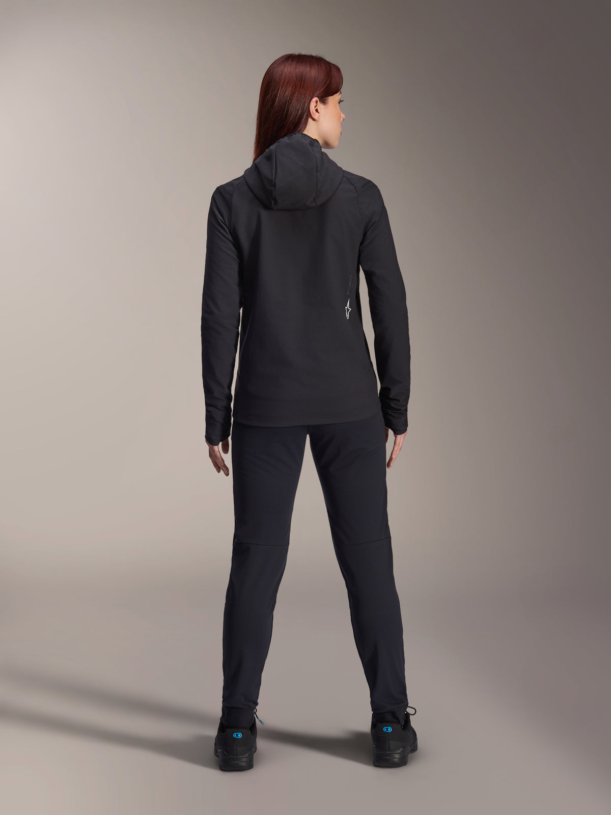 Woman wearing Alpinestars Stella A-Dura Thermal Jacket, back view, black insulated cycling jacket with hood, matching black pants and shoes, featuring a small white logo on the lower back