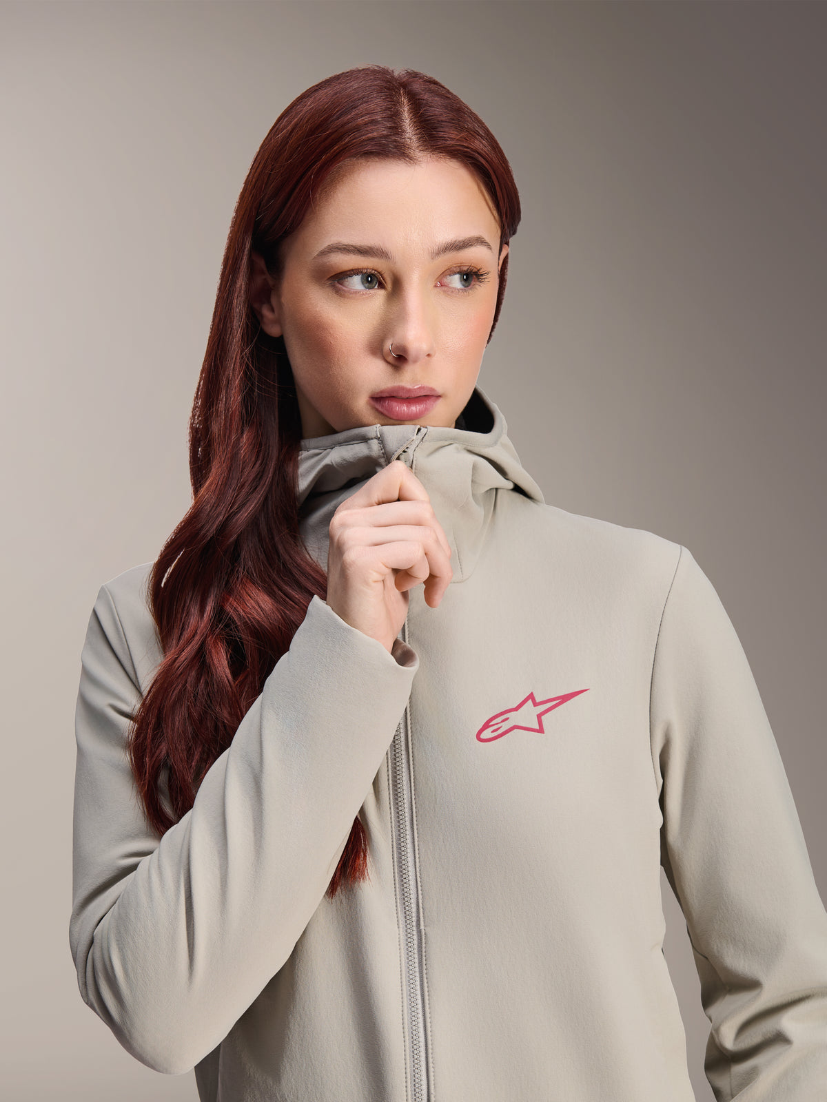 Alpinestars Stella A-Dura Thermojacke für Damen, Titanium-Grau mit pinkfarbenem Alpinestars Stern-Logo, Modell mit langem rotem Haar trägt die Kapuzenjacke und schließt den Reißverschluss, leichte isolierte Radbekleidung für kaltes Wetter