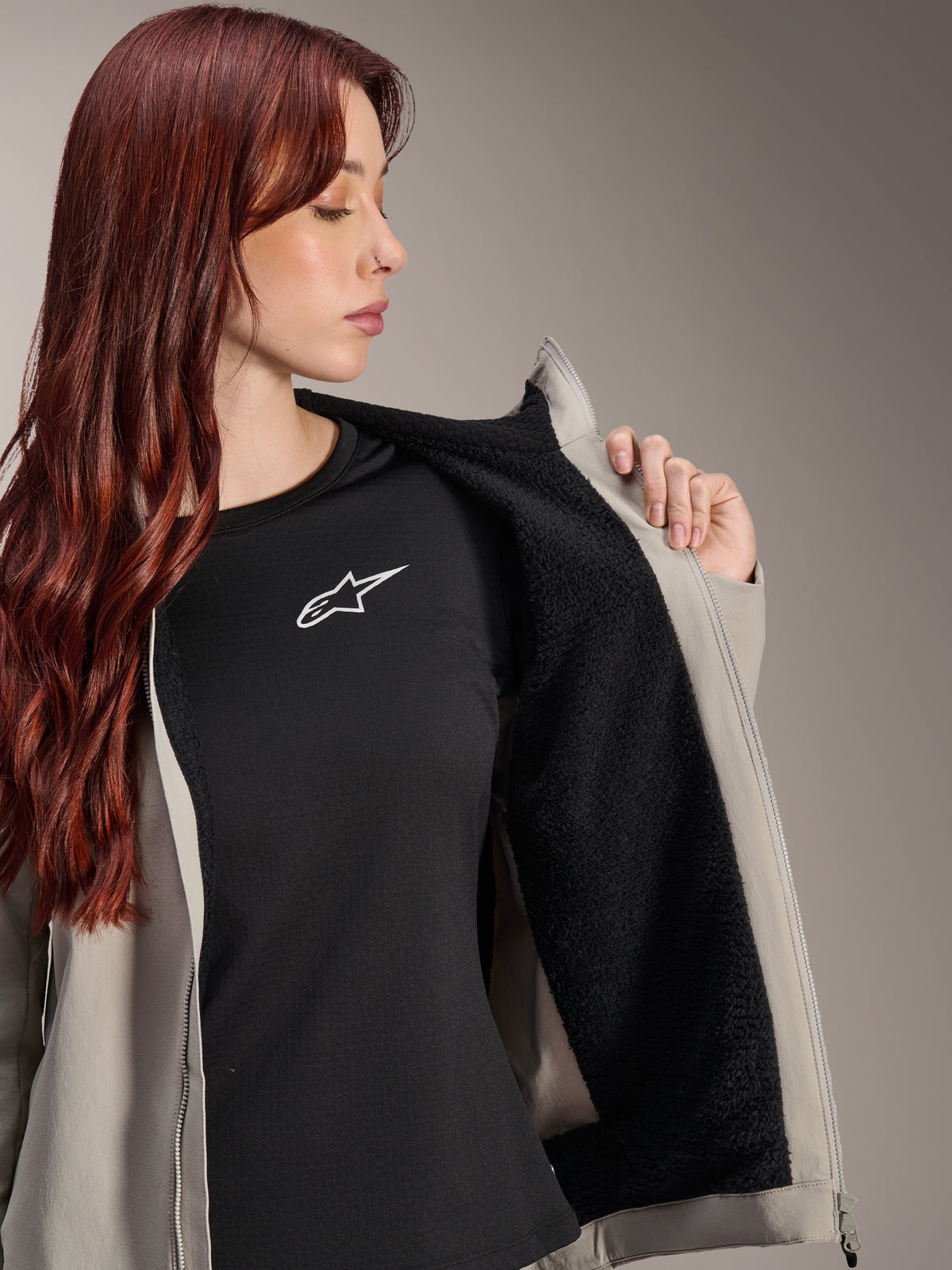 Frau in der Alpinestars Stella A-Dura Thermal Jacke in Titanium, zeigt das schwarze, strukturierte Fleece-Futter, getragen über einem schwarzen T-Shirt mit weißem Stern-Logo, leichte Mountainbike-Jacke für kalte Bedingungen.