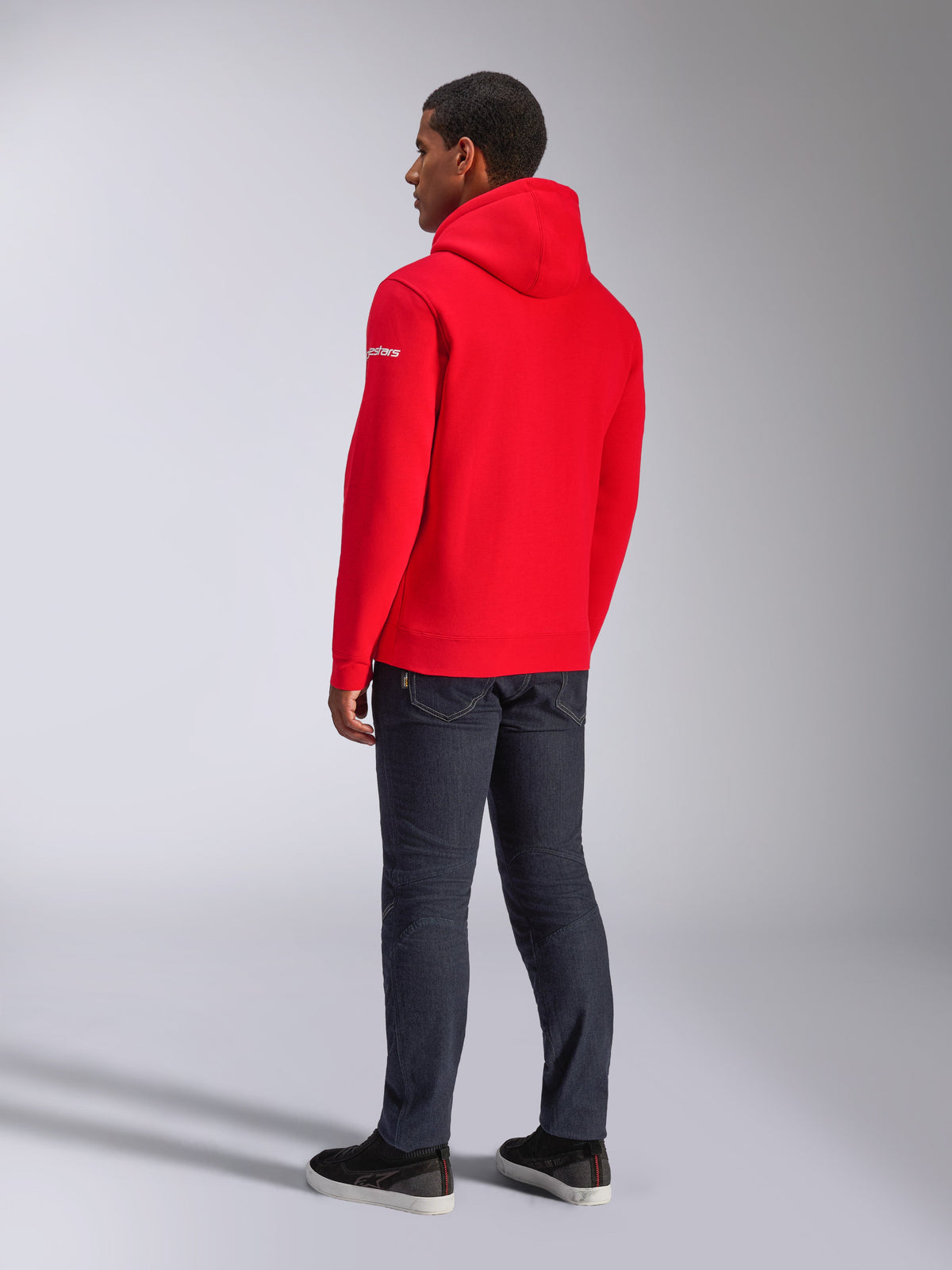 Alpinestars Frontal Hoodie, roter Pullover-Fleece-Hoodie, Rückansicht mit weißem Wortmarken-Logo auf dem Ärmel, getragen mit dunkler Jeanshose und schwarzen Schuhen, lässige Motorsport-Streetwear