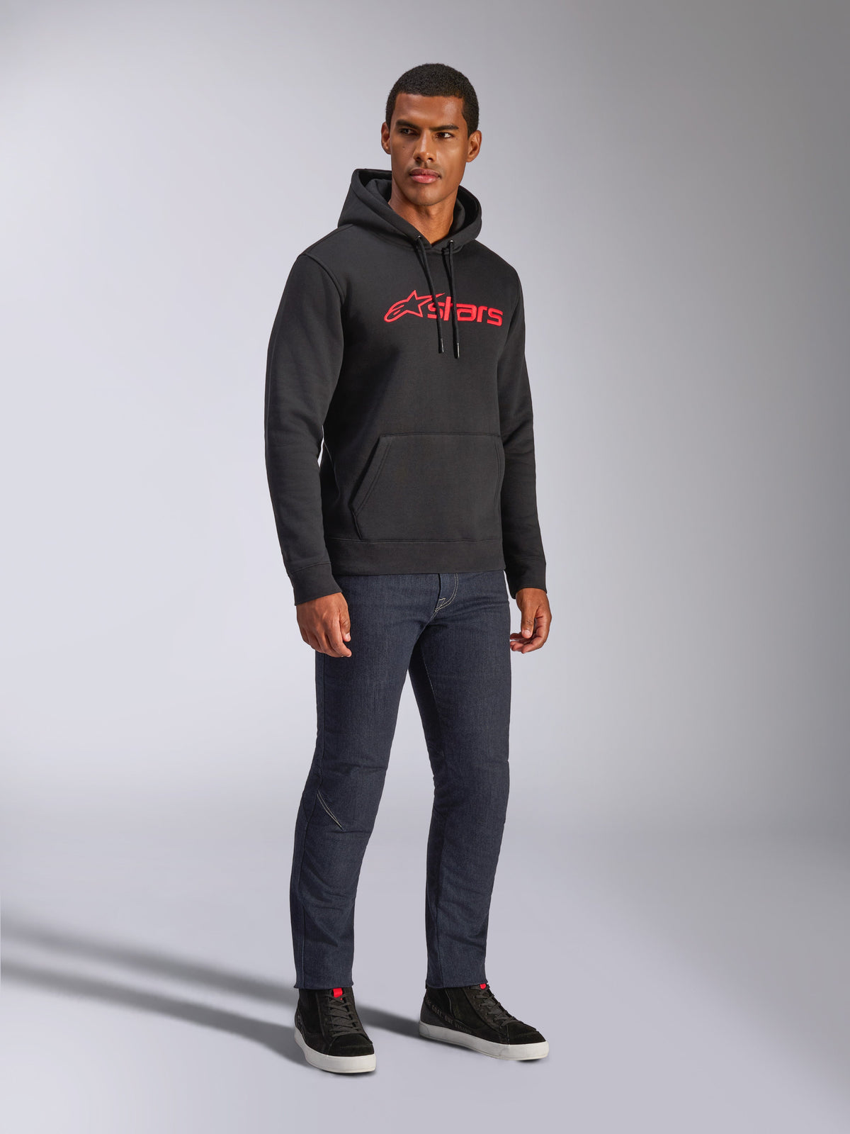 Alpinestars Blaze V3 Hoodie, black pullover fleece featuring a red Alpinestars logo on the chest, worn by a male model with dark denim jeans and black sneakers, casual motorsport-inspired streetwear