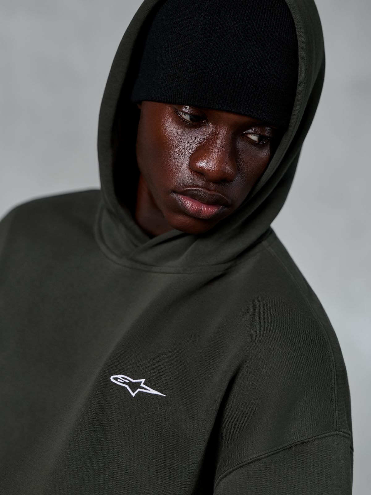 Alpinestars Essentials Ageless Hoodie, Off Black, close-up of model wearing a black beanie under the hood, featuring a small white star logo on the chest, casual motorsport-inspired streetwear