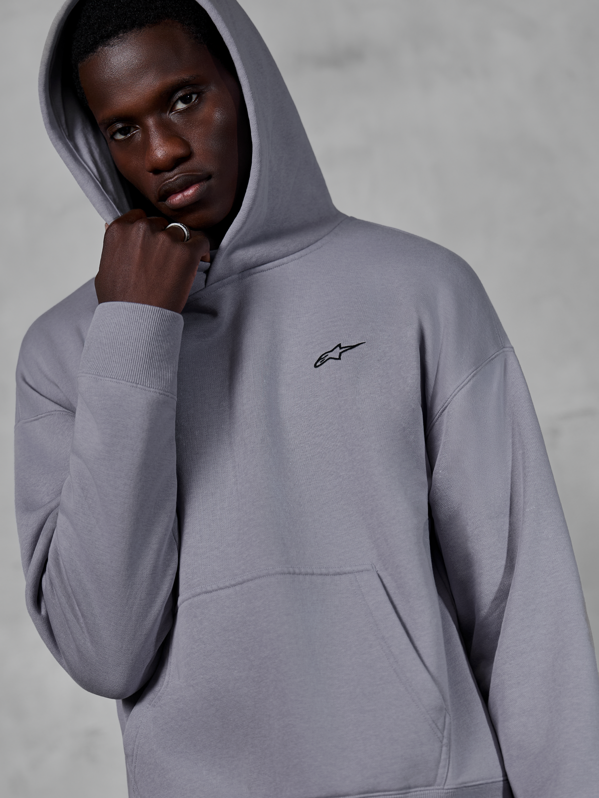 Alpinestars Essentials Ageless Hoodie, Silber, männliches Model trägt den Kapuzenpullover mit aufgesetzter Kapuze, kleines schwarzes Alpinestars-Logo auf der Brust, Kängurutasche, Rippbündchen, lässige Lifestyle-Streetwear