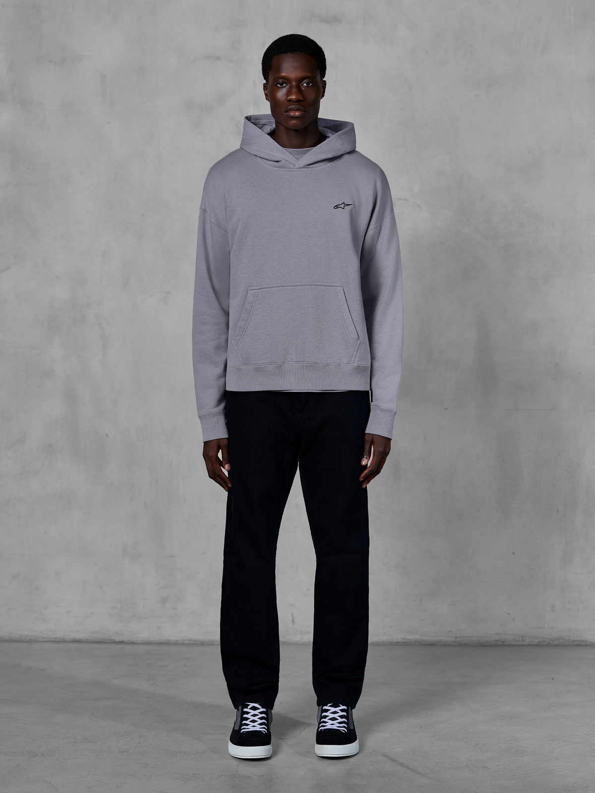 Alpinestars Essentials Ageless Hoodie, Silber, männliches Model in lässiger Streetwear, kleines schwarzes Alpinestars-Logo auf der Brust, Kängurutasche vorne, minimalistisches Design, kombiniert mit schwarzer Hose und Sneakern.