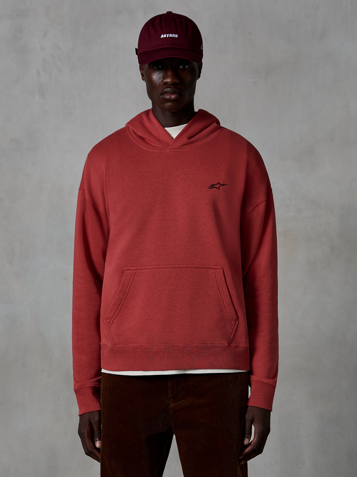 Alpinestars Essentials Ageless Hoodie, lässige Streetwear, Ziegelrot, mit kleinem schwarzen Stern-Logo auf der Brust, Kängurutasche, Stone-Wash-Finish, getragen von einem Model mit weinroter Kappe und brauner Cordhose