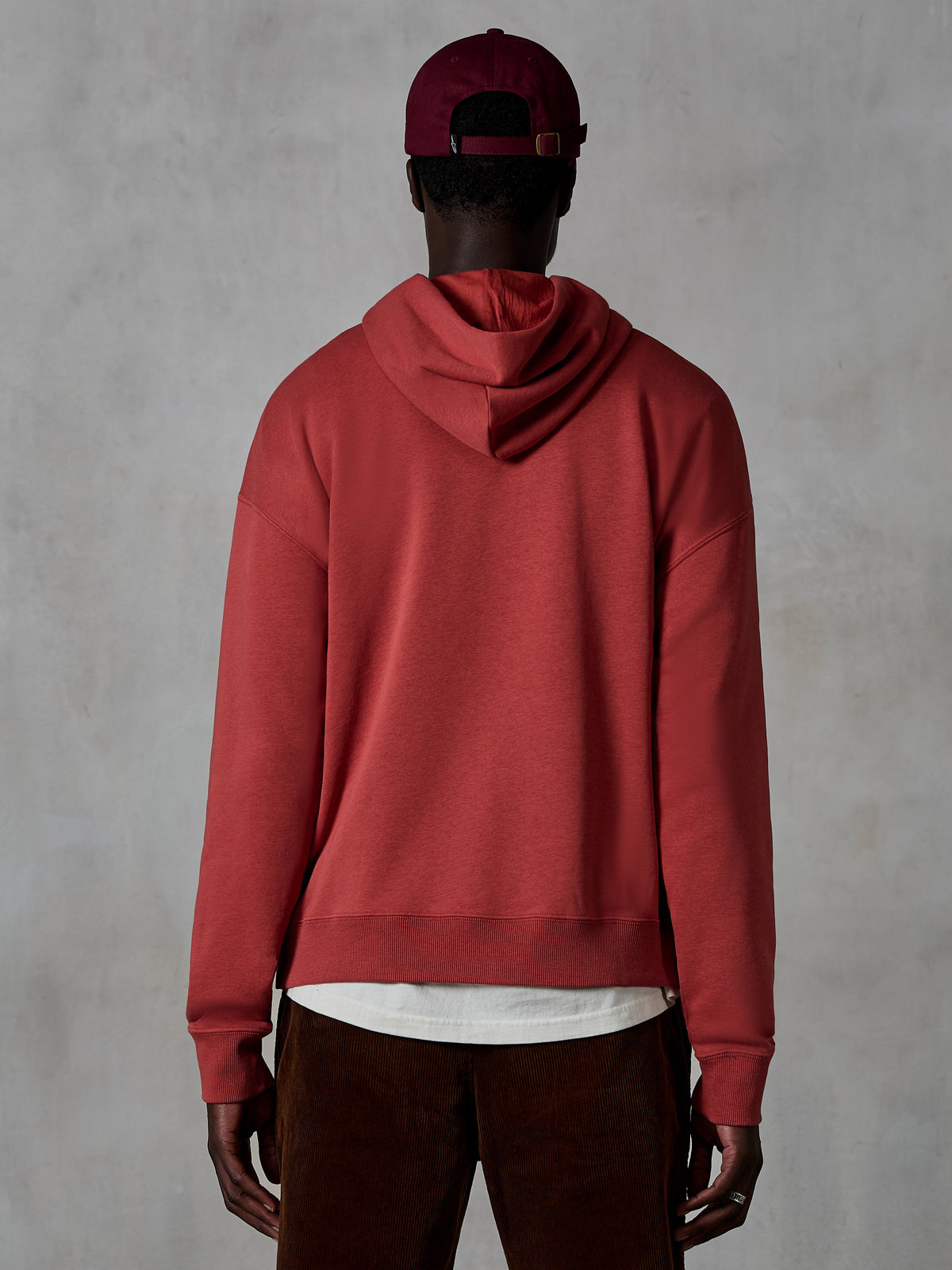 Alpinestars Essentials Ageless Hoodie, lässiger Fleece-Hoodie, Ziegelrot, Rückansicht eines Models in Lifestyle-Bekleidung, klares Design mit geripptem Saum und Bündchen, kombiniert mit einer braunen Cordhose und einer burgunderroten Kappe