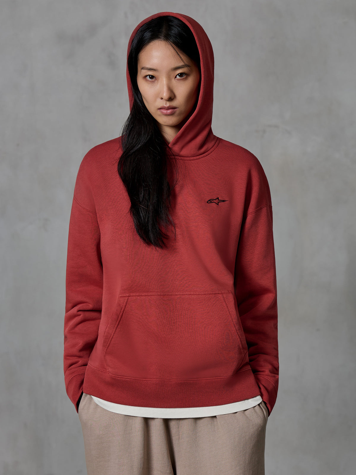 Alpinestars Essentials Ageless Hoodie, ziegelroter Kapuzenpullover, getragen von einem weiblichen Model mit aufgesetzter Kapuze, kleines schwarzes gesticktes Stern-Logo auf der Brust, Kängurutasche, lässige Motorsport-inspirierte Lifestyle-Streetwear.