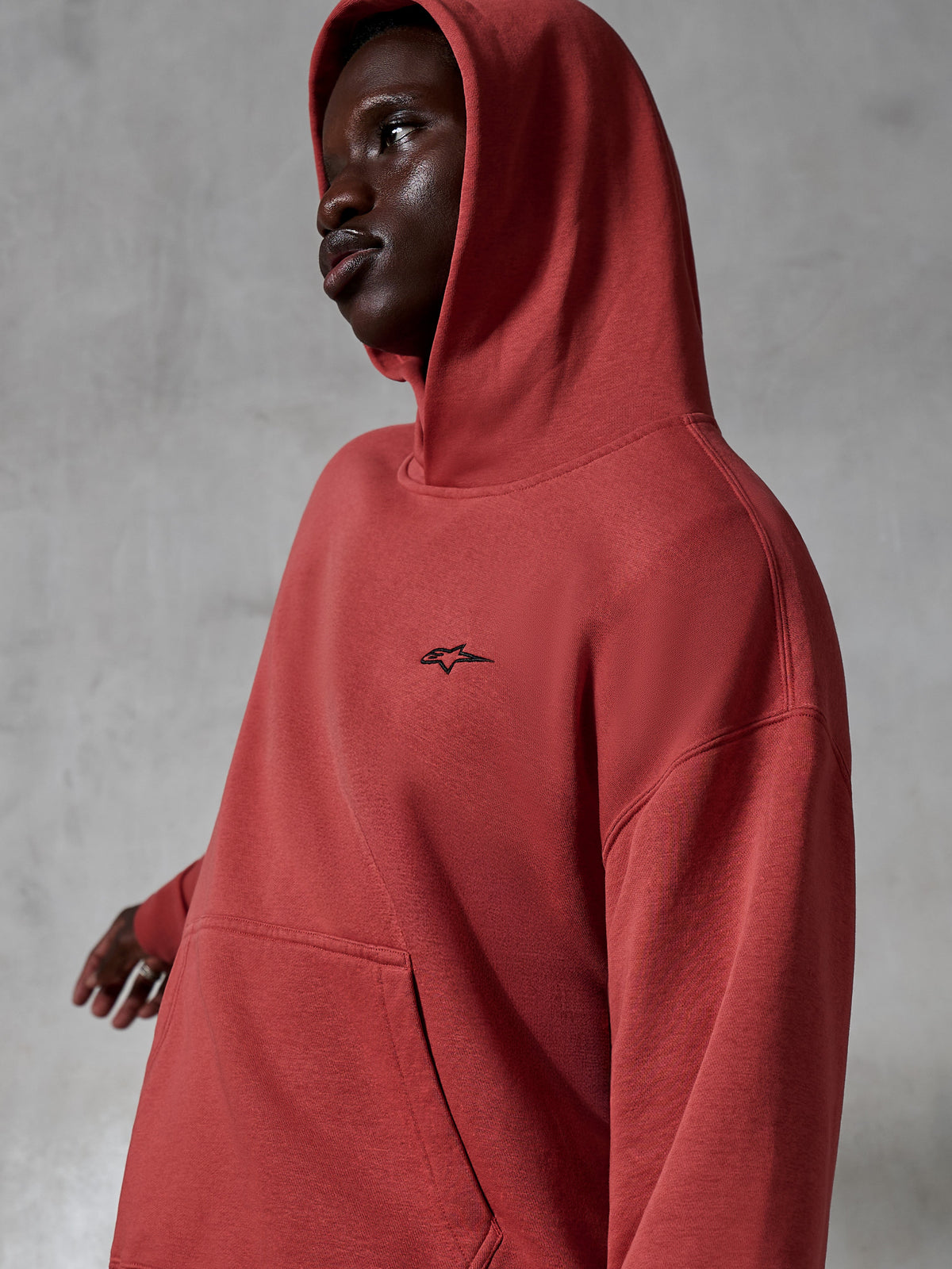 Alpinestars Essentials Ageless Hoodie, lässige Sportbekleidung, Ziegelrot, von einem Model mit aufgesetzter Kapuze getragen, mit einem kleinen schwarzen bestickten Logo, weiche Fleece-Textur, Streetwear mit entspannter Passform