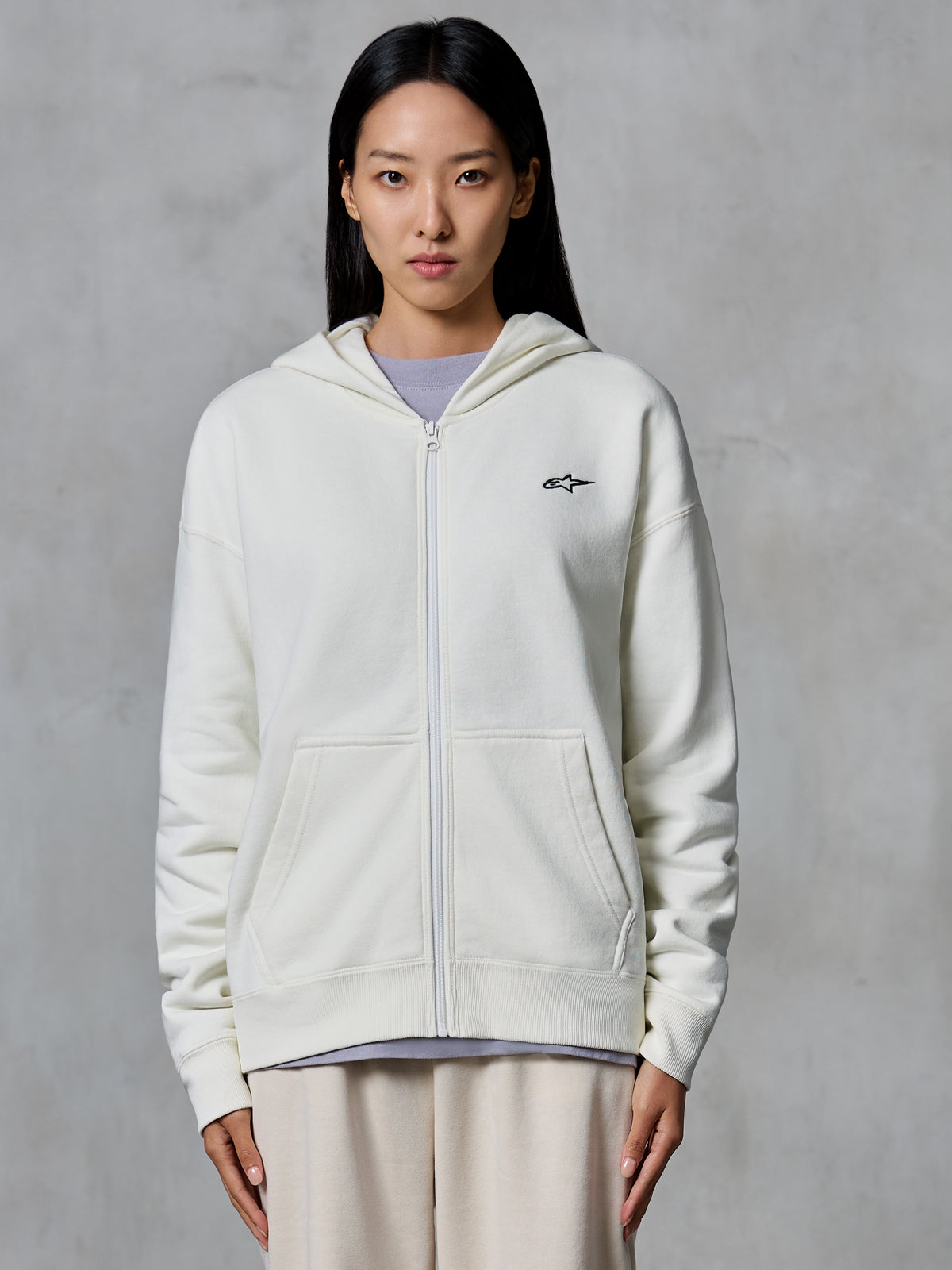 Alpinestars Essentials Ageless Zip Hoodie, Off-White, Fleece-Sweatjacke mit Reißverschluss vorne, von einem Model getragen, mit kleinem schwarzem Astars-Logo auf der Brust, entspannte Passform, lässige, vom Motorsport inspirierte Lifestyle-Bekleidung