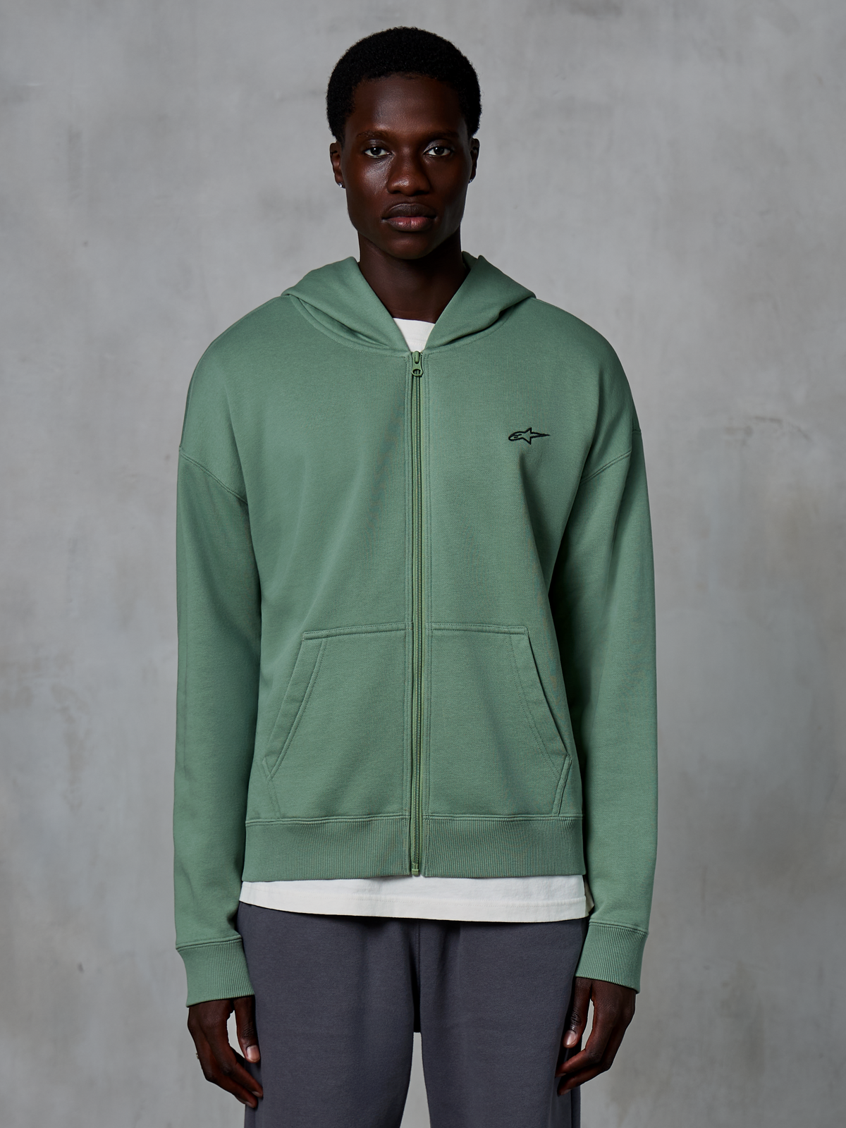 Alpinestars Essentials Ageless Zip Hoodie, lässige Streetwear-Kapuzenjacke, Kaktusgrün, mit dezentem gestickten Brustlogo, geteilten Kängurutaschen und Rippbündchen, getragen von einem männlichen Model vor einem strukturierten grauen Hintergrund