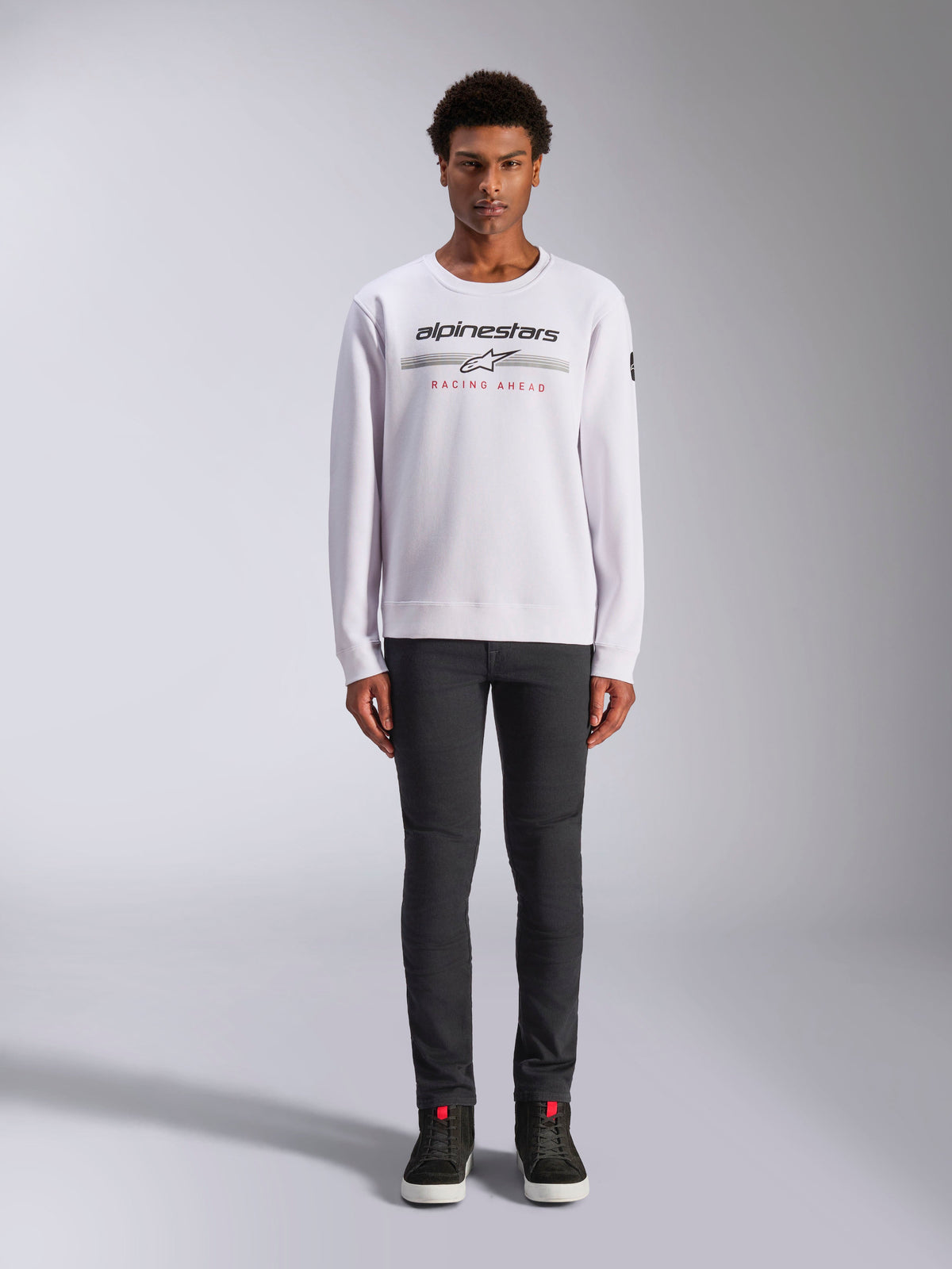 Alpinestars Bettern Crew, white crew neck sweatshirt, worn by male model with dark grey pants and black high-top sneakers, featuring Alpinestars branding with 'Racing Ahead' red text, casual motorsport-inspired streetwear