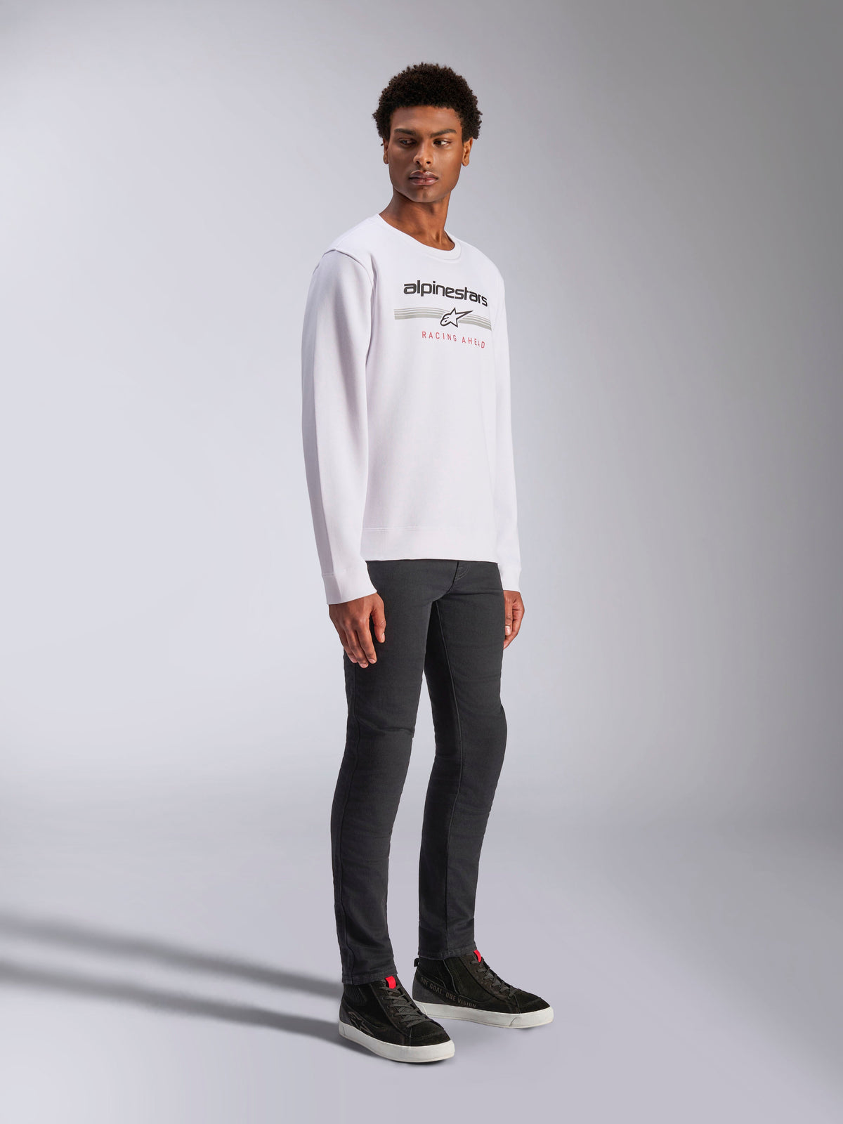 Alpinestars Bettern Crew, White Pullover Sweatshirt, featuring black Alpinestars logo and 'Racing Ahead' red text, casual motorsport streetwear, worn by a model with dark pants and black sneakers