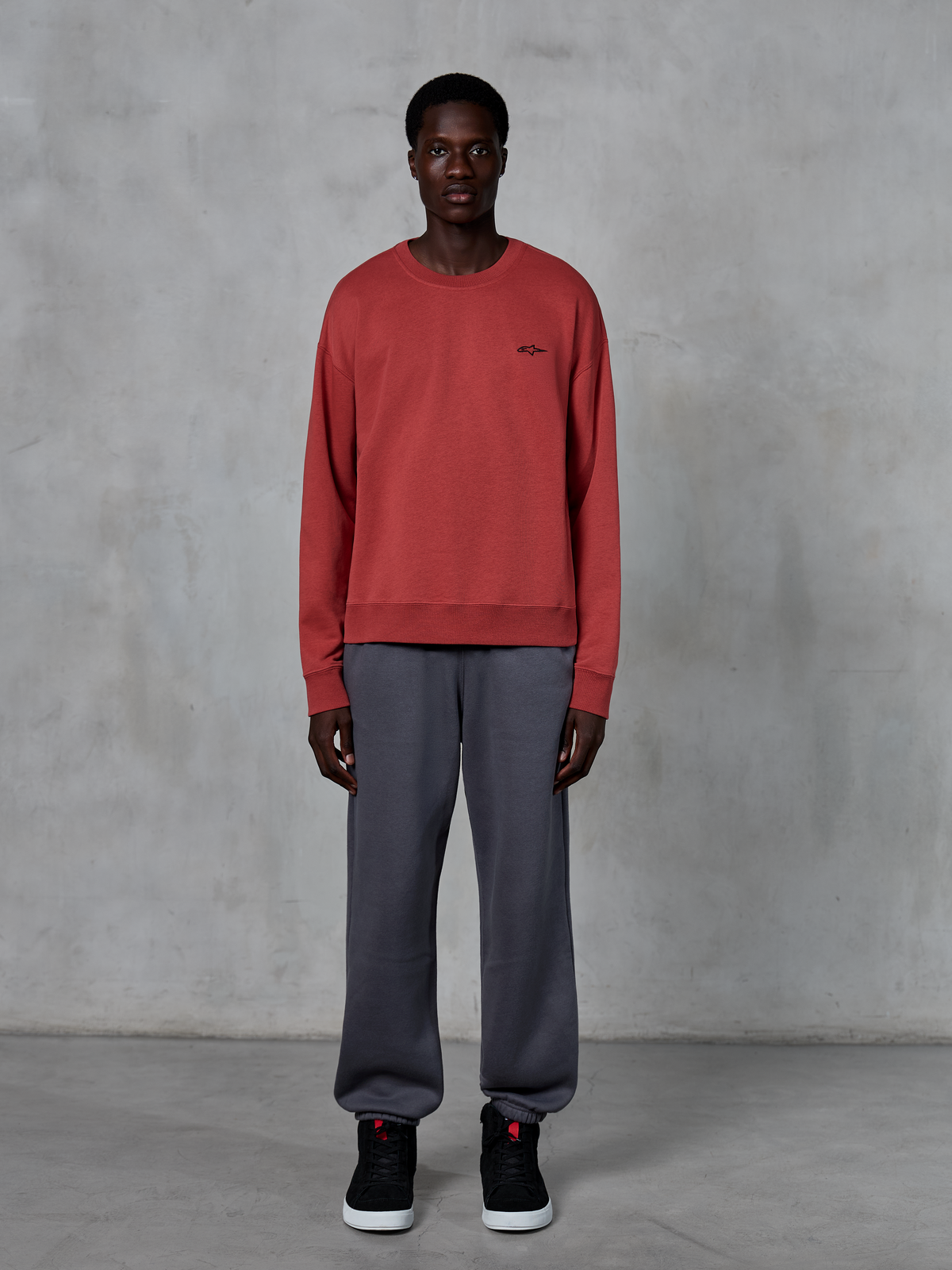 Alpinestars Essentials Ageless Crew, ziegelrotes Rundhals-Sweatshirt, getragen mit grauer Jogginghose und schwarzen Sneakern, mit einem kleinen schwarzen Logo auf der Brust, lässige, vom Motorsport inspirierte Streetwear