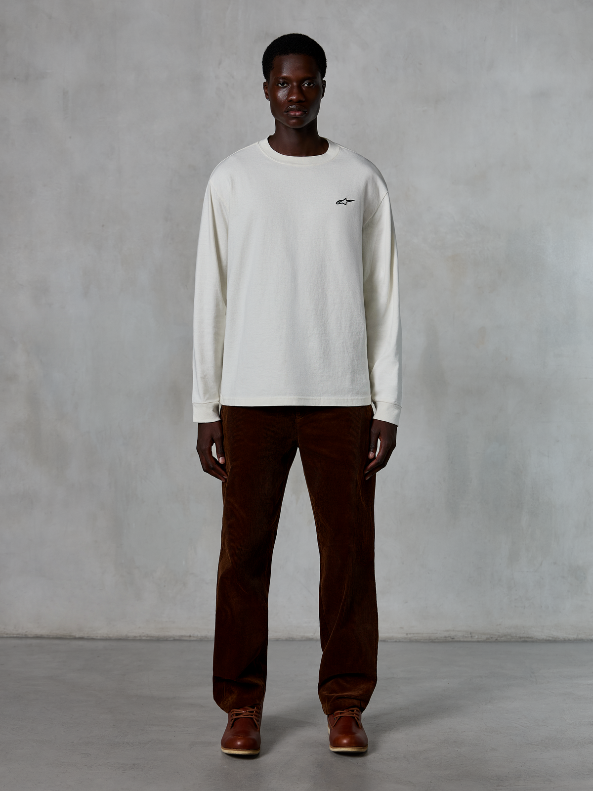 Alpinestars Essentials Ageless LS Tee, Langarmshirt, Off-White, getragen von einem Model mit brauner Cordhose und braunen Lederstiefeln, mit einem kleinen schwarzen Logo auf der Brust, entspannte zeitgemäße Passform, lässige Lifestyle-Streetwear