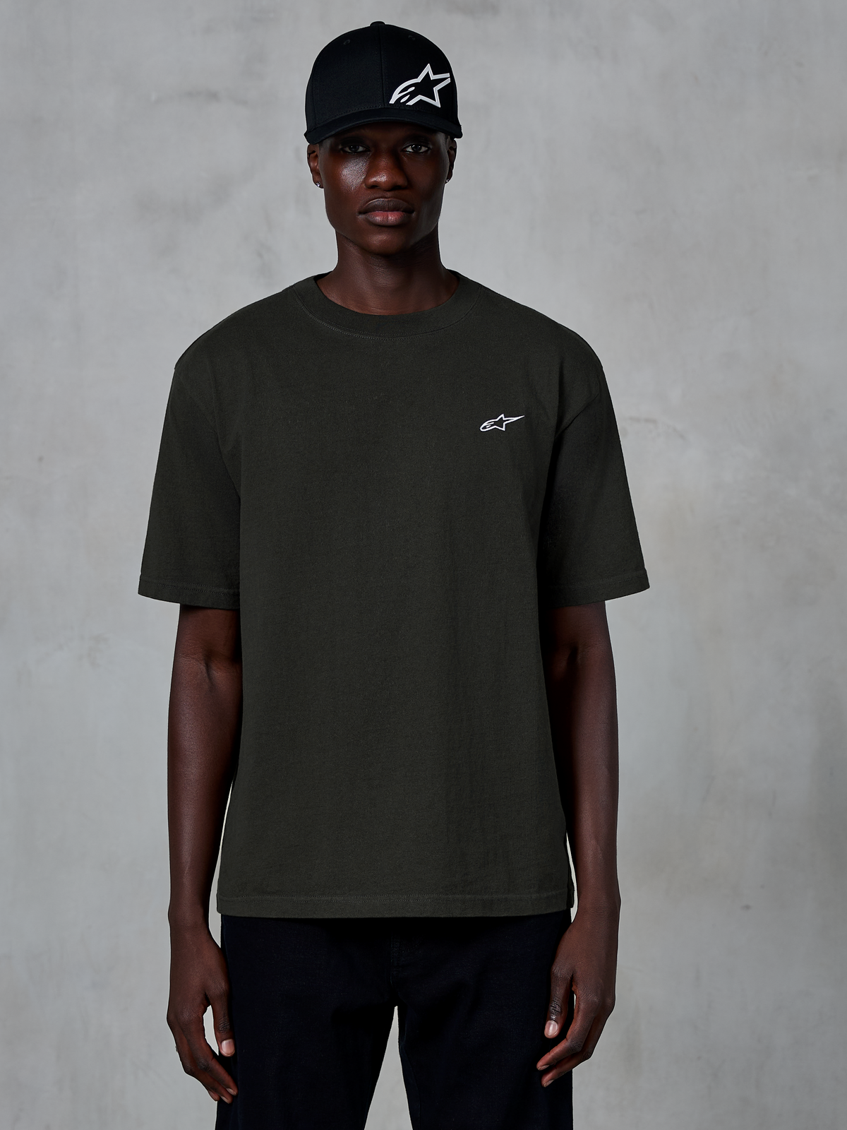 Alpinestars Essentials Ageless SS Tee, Kurzarm-T-Shirt, Off-Black, getragen von einem Modell mit einer schwarzen Logo-Kappe, kleines weißes Alpinestars-Logo auf der Brust, lässige vom Motorsport inspirierte Streetwear