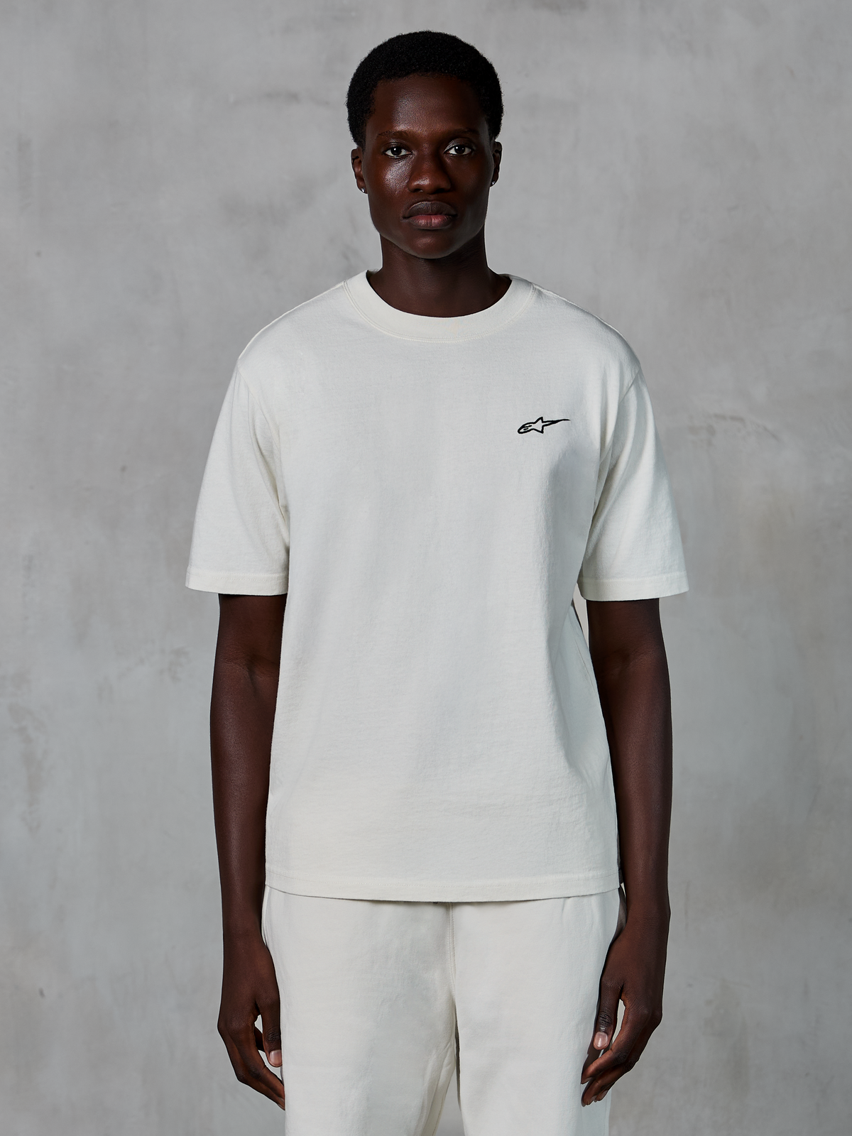 Alpinestars Essentials Ageless SS Tee, Kurzarm-T-Shirt, Off-White, von einem männlichen Model getragen, mit einem kleinen schwarzen Alpinestars-Logo auf der Brust, entspannte Passform, Bio-Baumwolljersey, lässige Motorsport-Streetwear