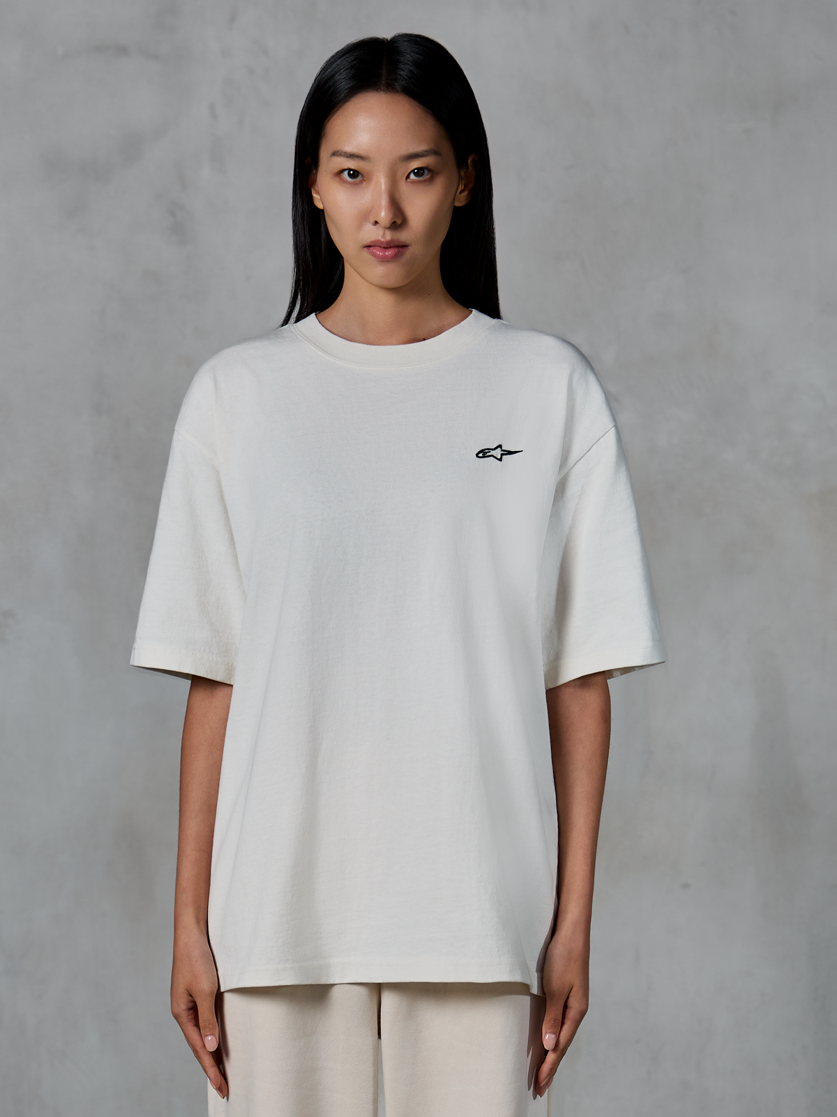 Alpinestars Essentials Ageless SS Tee, kurzärmeliges T-Shirt, Off-White, getragen von einem weiblichen Model, mit kleinem schwarzem Alpinestars-Logo auf der Brust, entspannte Passform, lässige Lifestyle-Streetwear