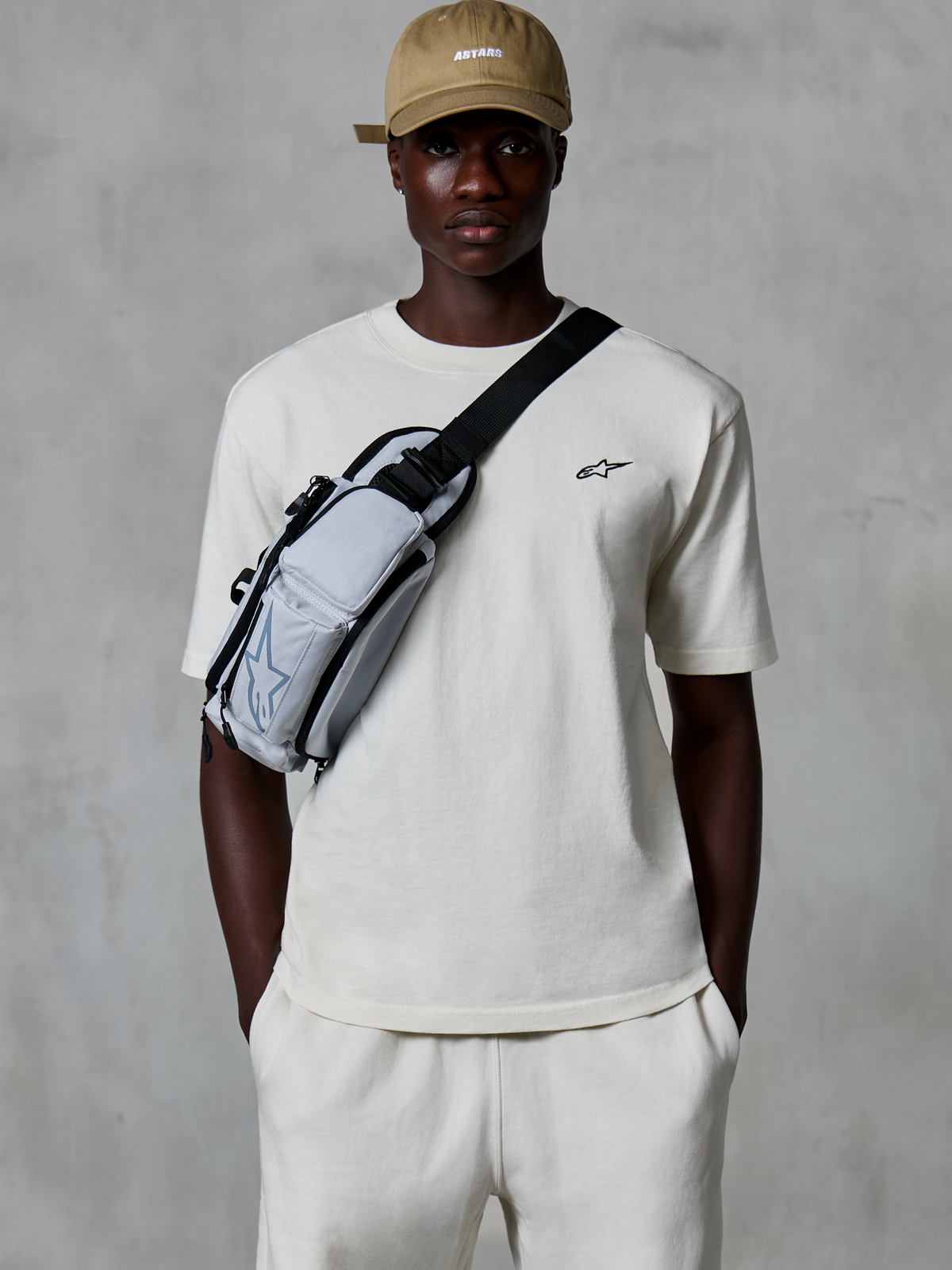 Alpinestars Essentials Ageless SS Tee, Kurzarm-T-Shirt, Off-White, getragen mit passenden Shorts und einer hellbraunen Kappe, mit einem kleinen schwarzen Logo auf der Brust und einer grauen Umhängetasche, lässige, vom Motorsport inspirierte Streetwear
