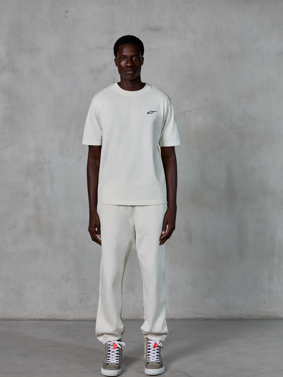Alpinestars Essentials Ageless SS Tee, kurzärmeliges T-Shirt, Off-White, mit kleinem schwarzen Alpinestars-Logo auf der Brust, kombiniert mit passender Jogginghose und Sneakern für einen entspannten Lifestyle-Look