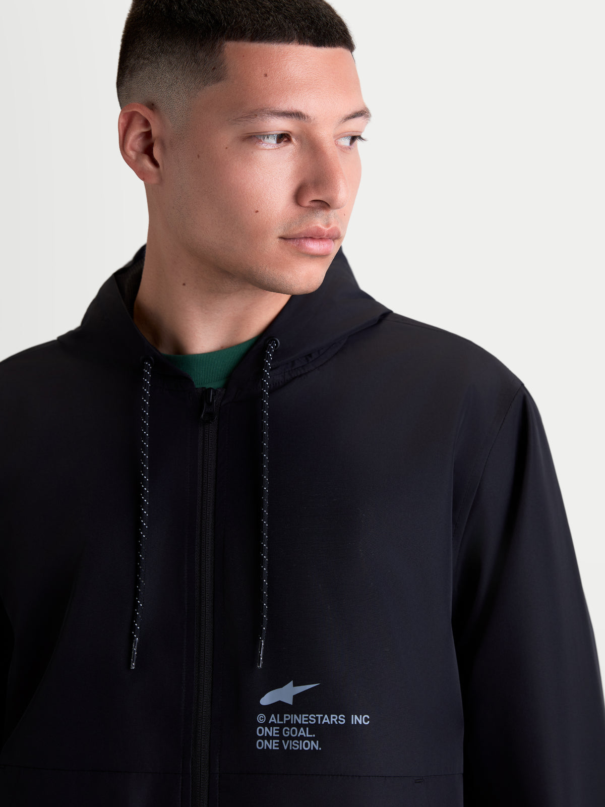 Alpinestars Speeder Resolve Winddichte Jacke, leichte Sportswear-Kapuzenjacke aus Polyester für Herren, Schwarz, Urban Layering, Detailansicht am Modell mit Alpinestars-Brustlogo und gesprenkelten Kordelzügen.