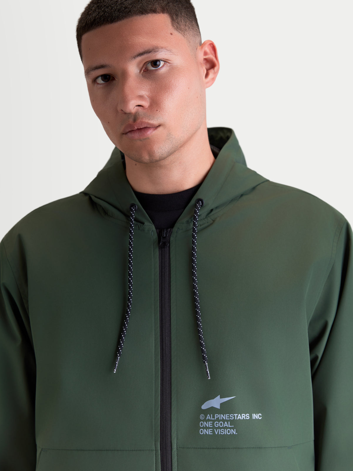 Alpinestars Speeder Resolve Winddichte Jacke, leichte Sportswear-Kapuzenjacke in Grün, mit verstellbarer Kapuze, gesprenkelten Kordelzügen und Alpinestars-Branding auf der Brust, getragen von einem männlichen Modell.