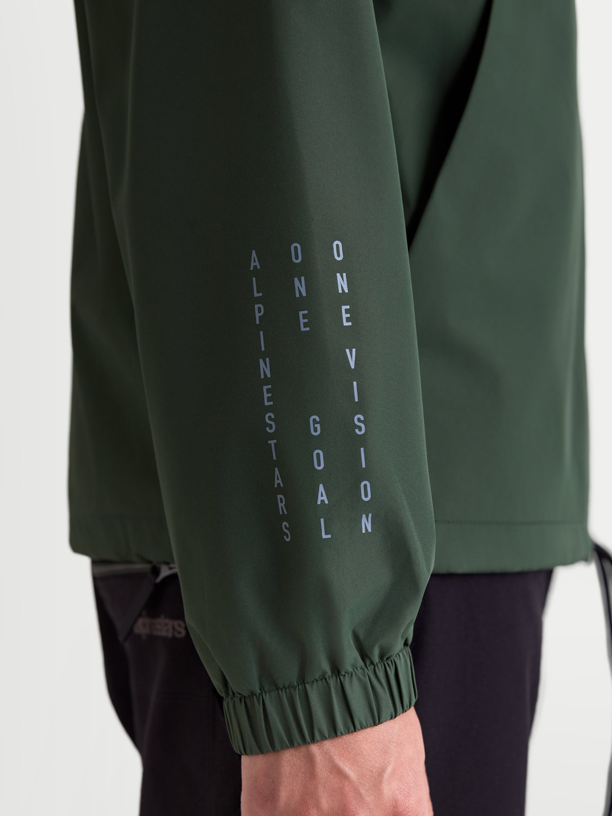 Alpinestars Speeder Resolve Winddichte Jacke, leichte Sportswear in Grün (Pine Green). Nahaufnahme des Ärmels mit vertikalem Alpinestars Branding, dem Motto „One Goal One Vision“ und einem elastischen Ärmelbündchen.