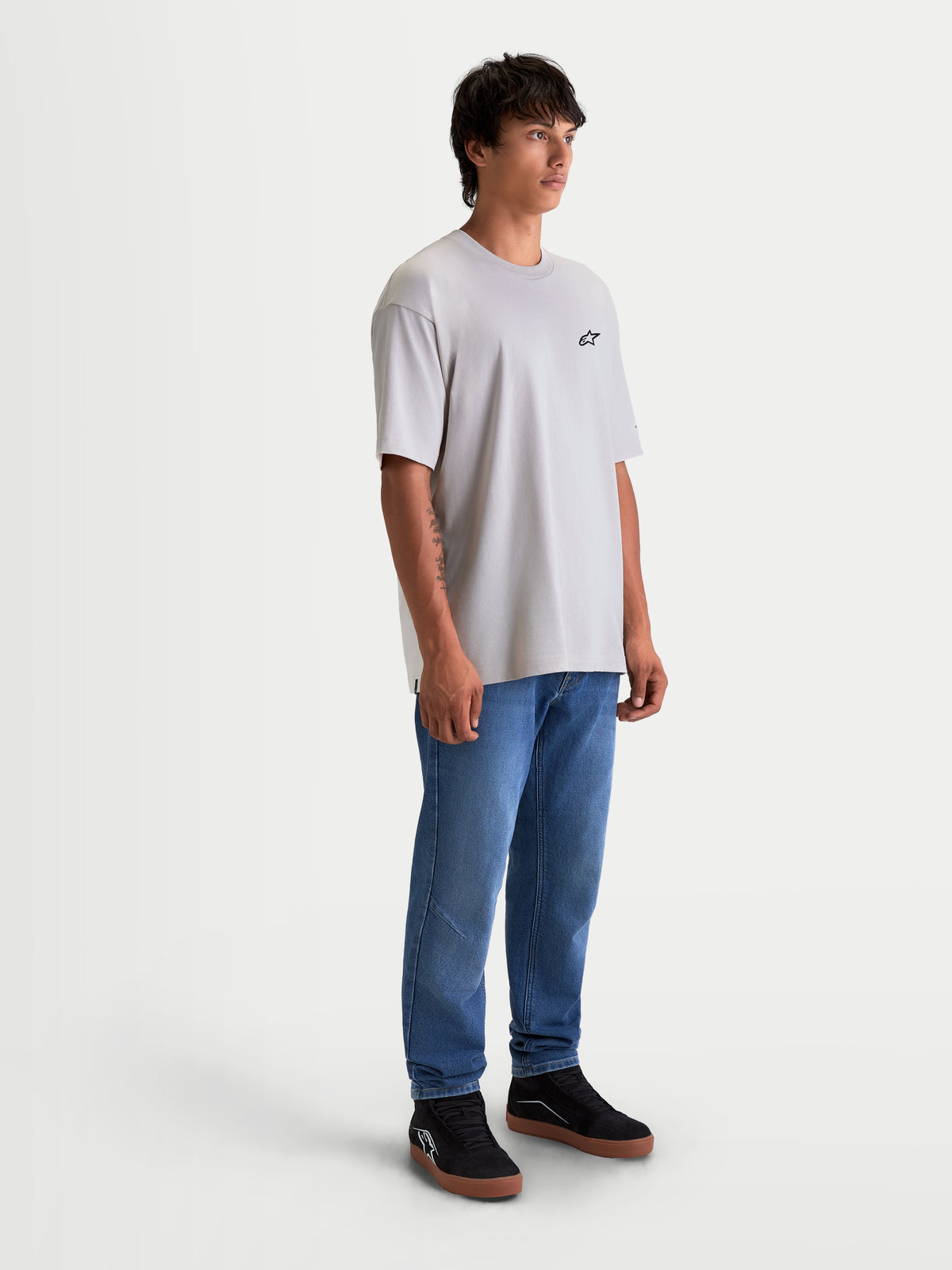 Alpinestars Mandate Oversized CSF T-Shirt, Kurzarm-Sportswear-T-Shirt, Unisex, Zementgrau, mit lässiger Oversized-Passform und kleinem schwarzen Astars-Logo auf der Brust. Hochwertiger Baumwoll-Polyester-Mix für alltäglichen Komfort. Gezeigt an einem männlichen Model mit blauen Jeans und schwarzen Sneakern.