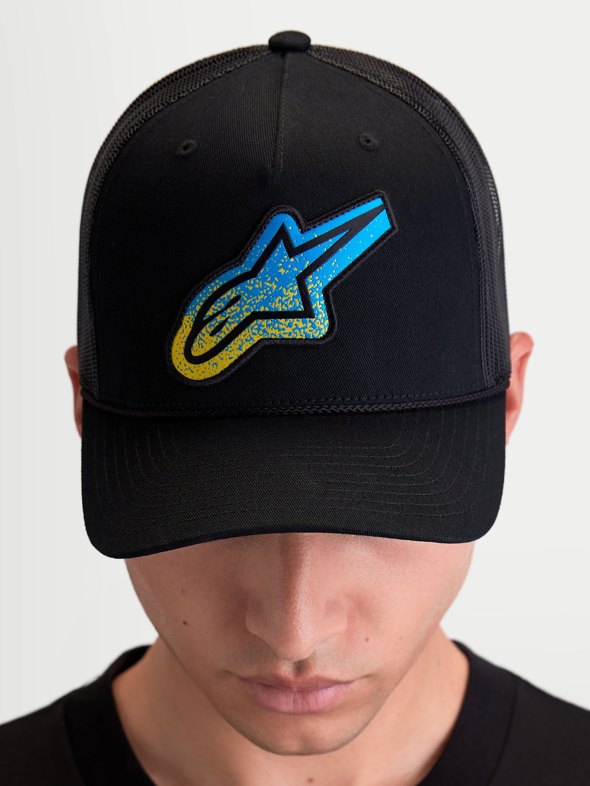 Alpinestars Meteorite Trucker-Kappe, Sportswear Trucker-Mütze mit Mesh-Rückseite, Schwarz und Blau, mit strukturiertem Astars Logo-Patch, strukturierter Krone und gebogenem Schirm, von einem Model getragen.