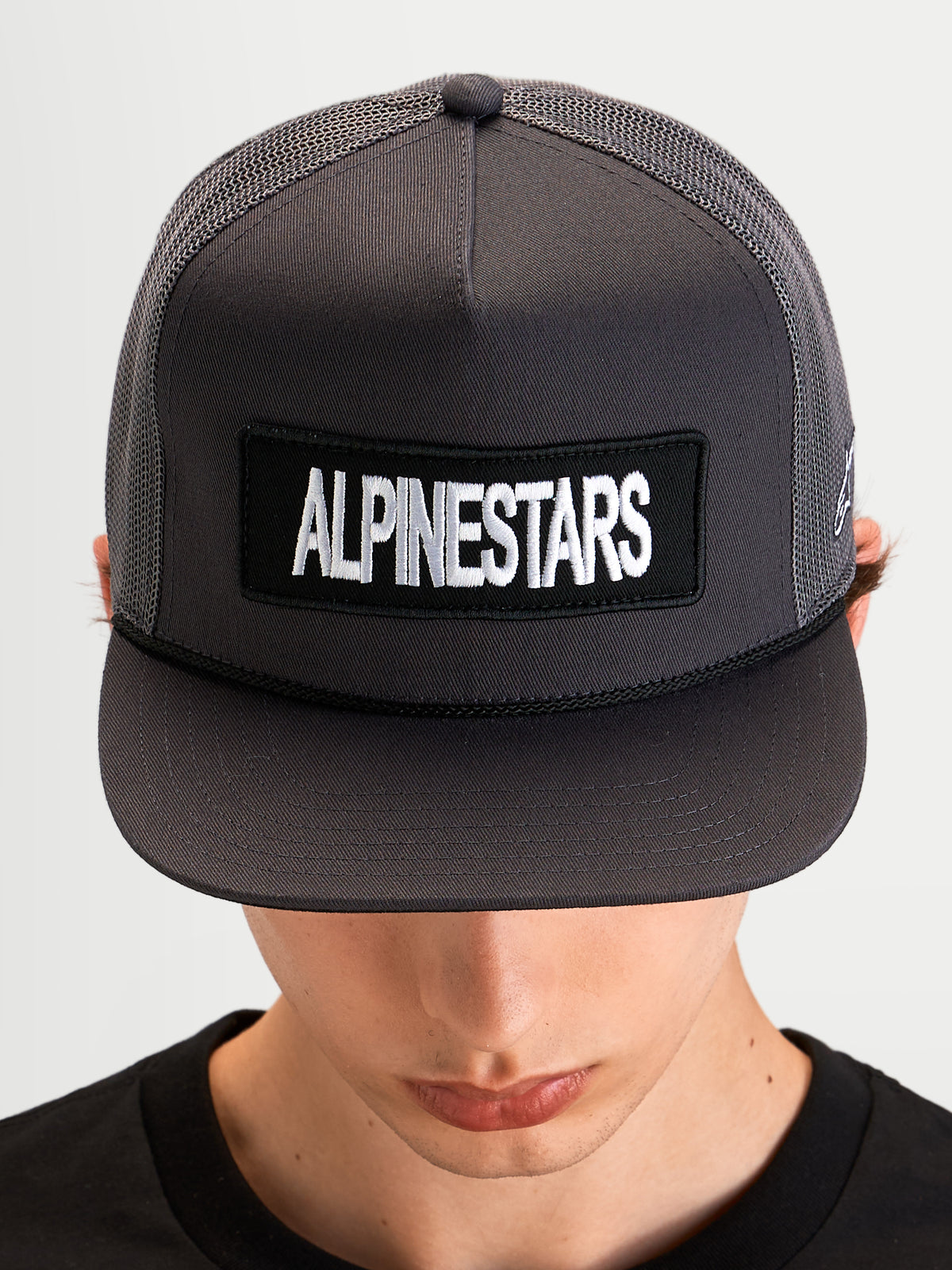 Alpinestars Ten Trucker-Kappe, flache Schirm-Cap für Sportswear in Dunkelgrau, getragen von einem Modell, mit strukturiertem Mesh-Trucker-Kopfbereich und Alpinestars Wortlogo-Patch auf der Vorderseite