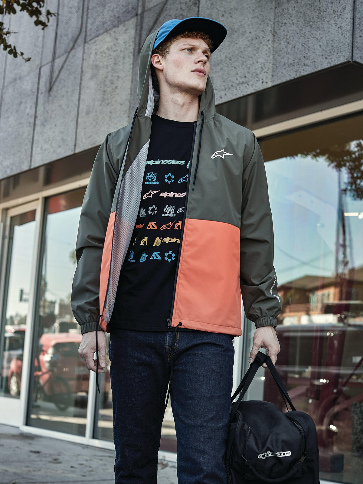 Man wearing Alpinestars Speeder Windbreaker Jacket in Military Orange, featuring a military green and orange color-blocked design, hooded with mesh lining, paired with a black graphic logo t-shirt, blue jeans, and carrying a black Alpinestars duffel bag, casual sportswear lifestyle look