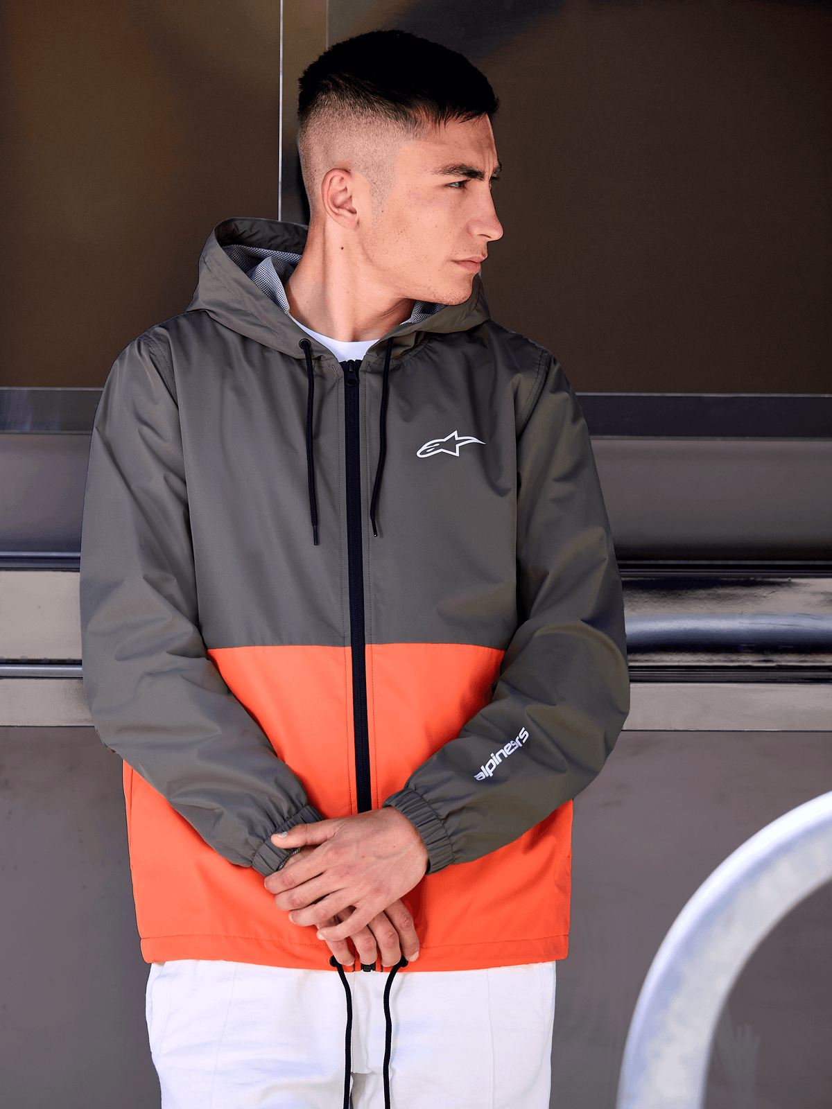 Man wearing the Alpinestars Speeder Windbreaker Jacket, Military Orange and Dark Green color-blocked design, hooded casual outerwear with white logos on chest and sleeve, paired with white pants for a motorsport-inspired lifestyle look