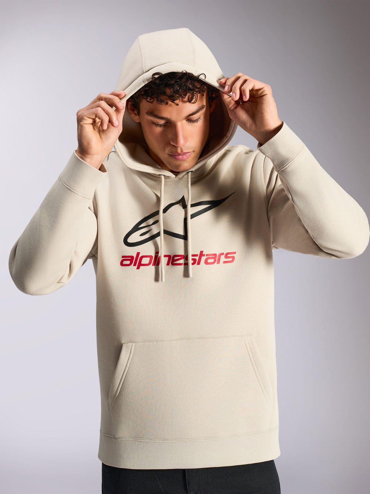 Alpinestars Always V3 Hoodie, männliches Modell in naturweißem Fleece-Kapuzenpullover, schwarzes und rotes lineares Logo auf der Vorderseite, Kapuze mit Kordelzug, Kängurutasche, lässige vom Motorsport inspirierte Streetwear