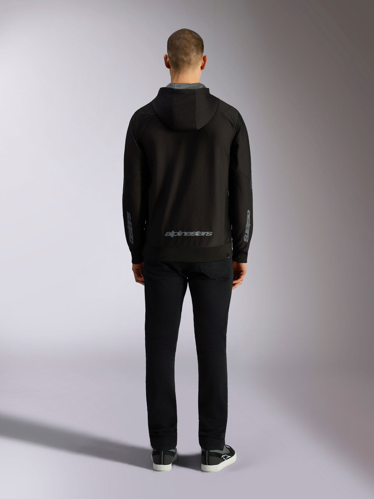 Rückansicht des Alpinestars Sessions V3 Hoodie in Schwarz-Grau, mit einer grauen Alpinestars-Wortmarke am unteren Rücken und Astars-Logos auf den Unterarmen, getragen von einem Model mit schwarzer Hose, lässige vom Motorsport inspirierte Streetwear
