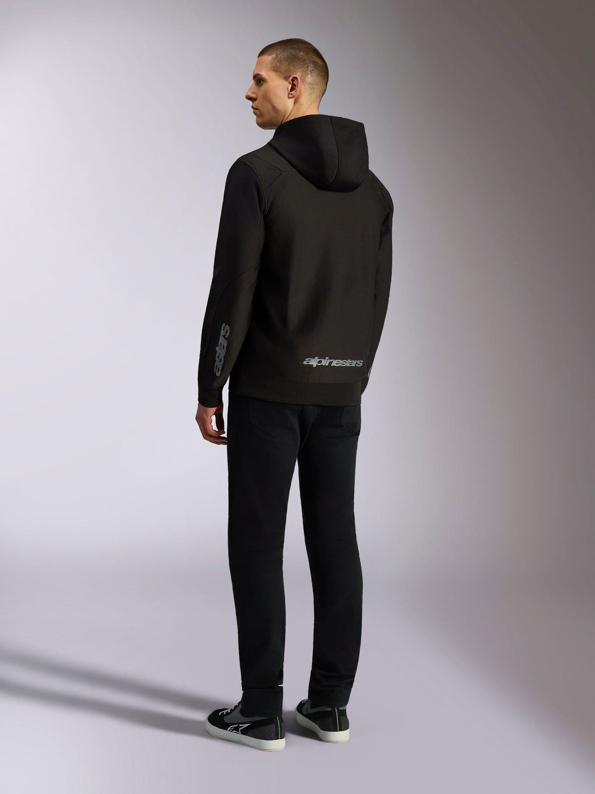 Alpinestars Sessions V3 Hoodie, schwarze Casual-Streetwear, Rückansicht an einem Model, mit grauem Alpinestars-Schriftzug am unteren Rücken und Astars-Logo auf dem Ärmel, Kapuzenjacke mit Reißverschluss und modernem, vom Motorsport inspiriertem Design