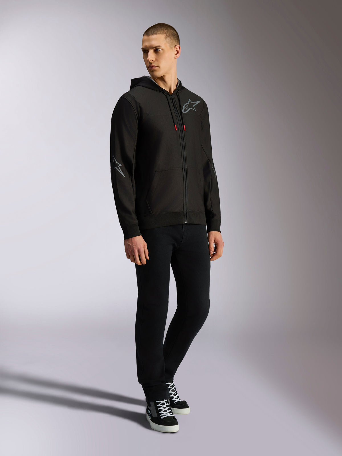 Alpinestars Sessions V3 Hoodie, Kapuzenjacke mit Front-Reißverschluss aus Polyfleece, Schwarz und Grau, graue Logos auf Brust und Ärmeln, Kapuze mit Kordelzug und roten Kordelenden, Ganzkörperansicht eines Models in lässiger Motorsport-Streetwear