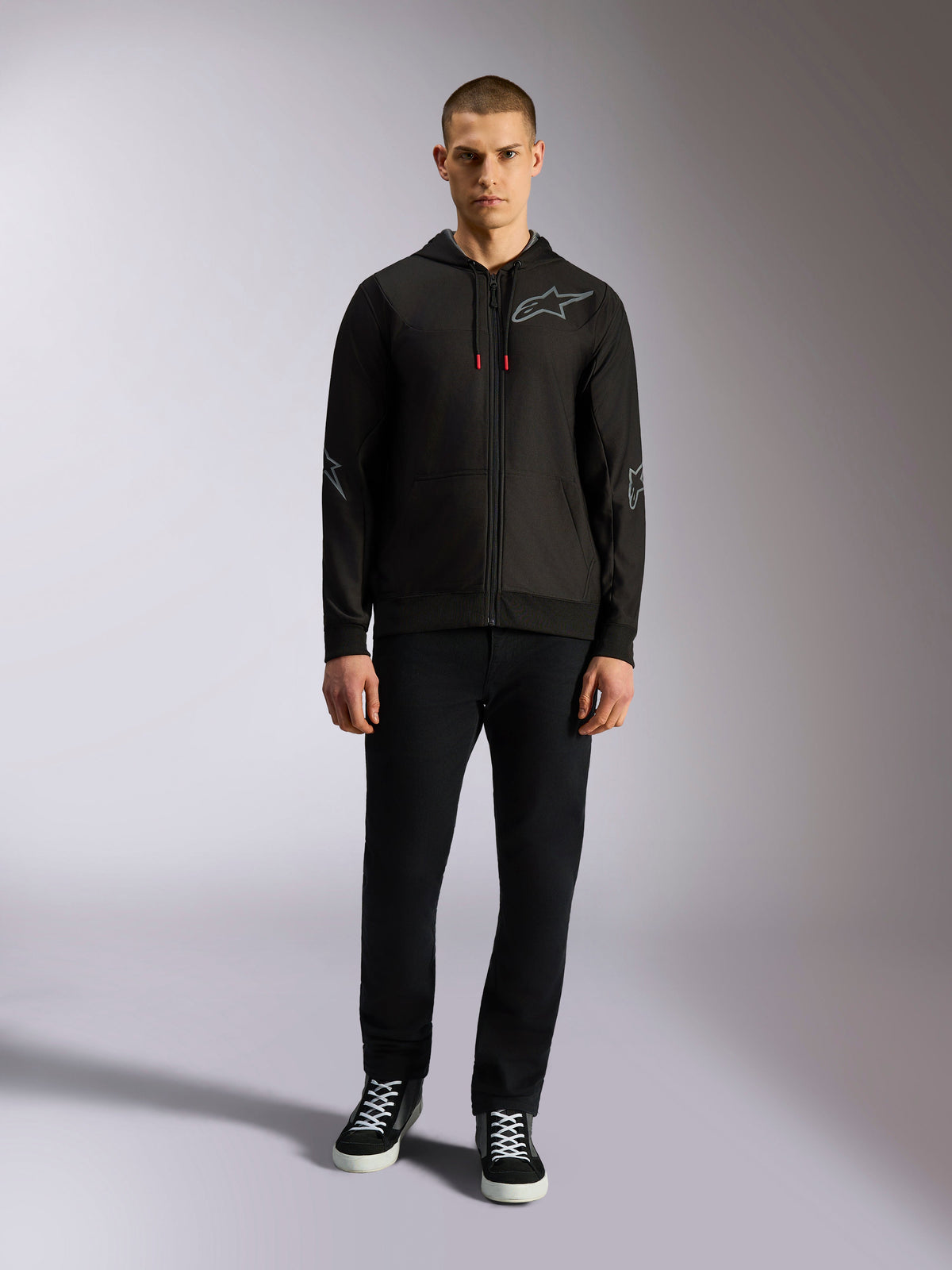 Alpinestars Sessions V3 Hoodie, schwarze Kapuzenjacke mit durchgehendem Reißverschluss, graues Astars-Brustlogo, Ellbogen- und Unterarmgrafiken, rote Kordelzug-Akzente, getragen von einem männlichen Model mit schwarzer Hose und Sneakern, lässige Motorsportbekleidung