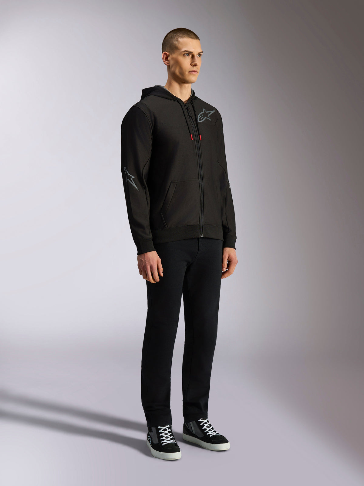 Alpinestars Sessions V3 Hoodie, lässige Motorsport-Streetwear, schwarze Kapuzenjacke mit Reißverschluss, grauem Stern-Logo auf Brust und Ärmel, Kordelzügen mit roten Enden, Rippbündchen und Saum, getragen von einem Model mit schwarzer Hose und Sneakern.