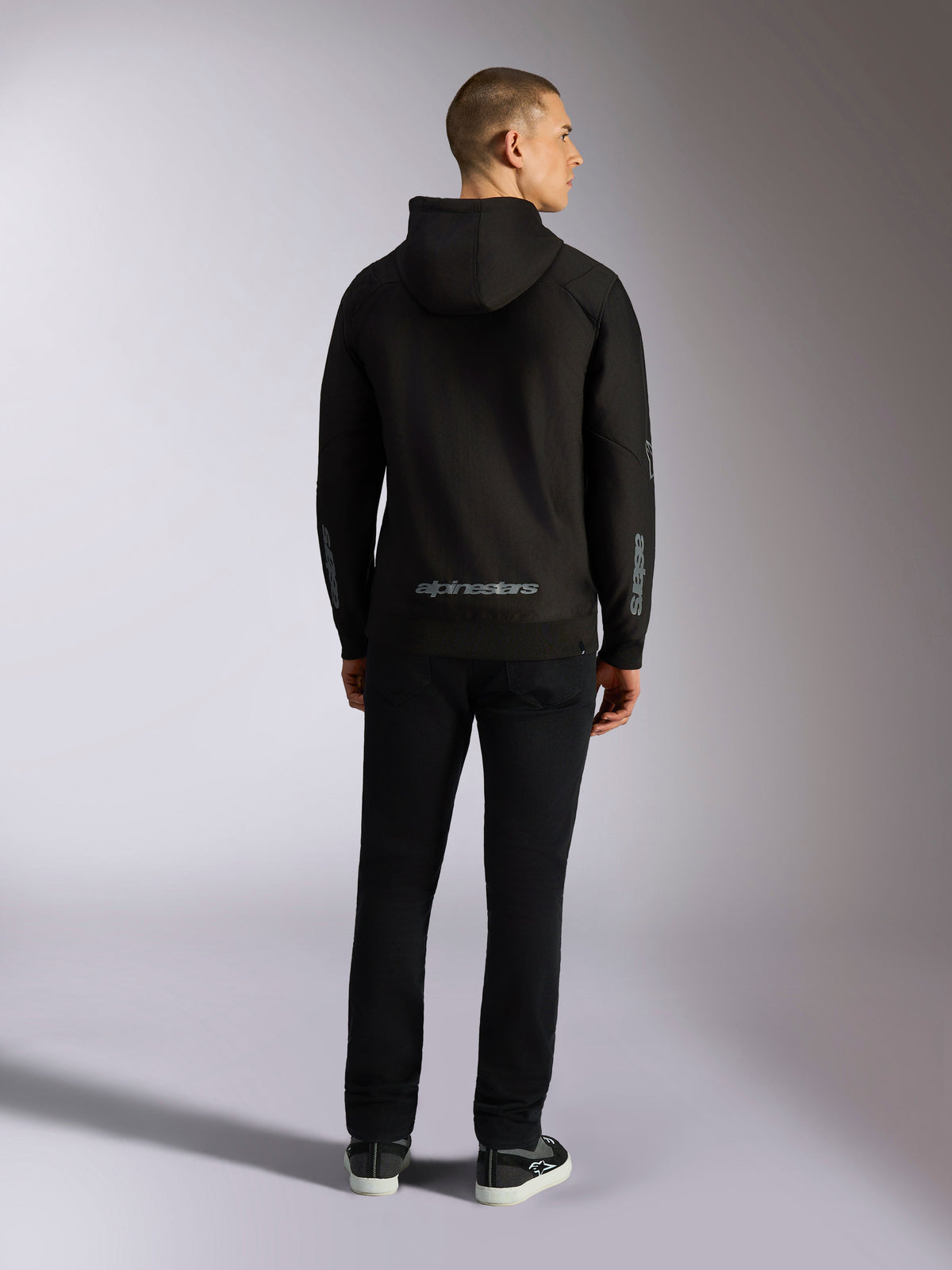 Alpinestars Sessions V3 Hoodie, lässige Streetwear, Schwarz mit grauem Alpinestars-Schriftzug am unteren Rücken und „astars“-Branding auf den Ärmeln, Rückansicht eines männlichen Models in Hoodie und schwarzer Hose, modernes, vom Motorsport inspiriertes Design