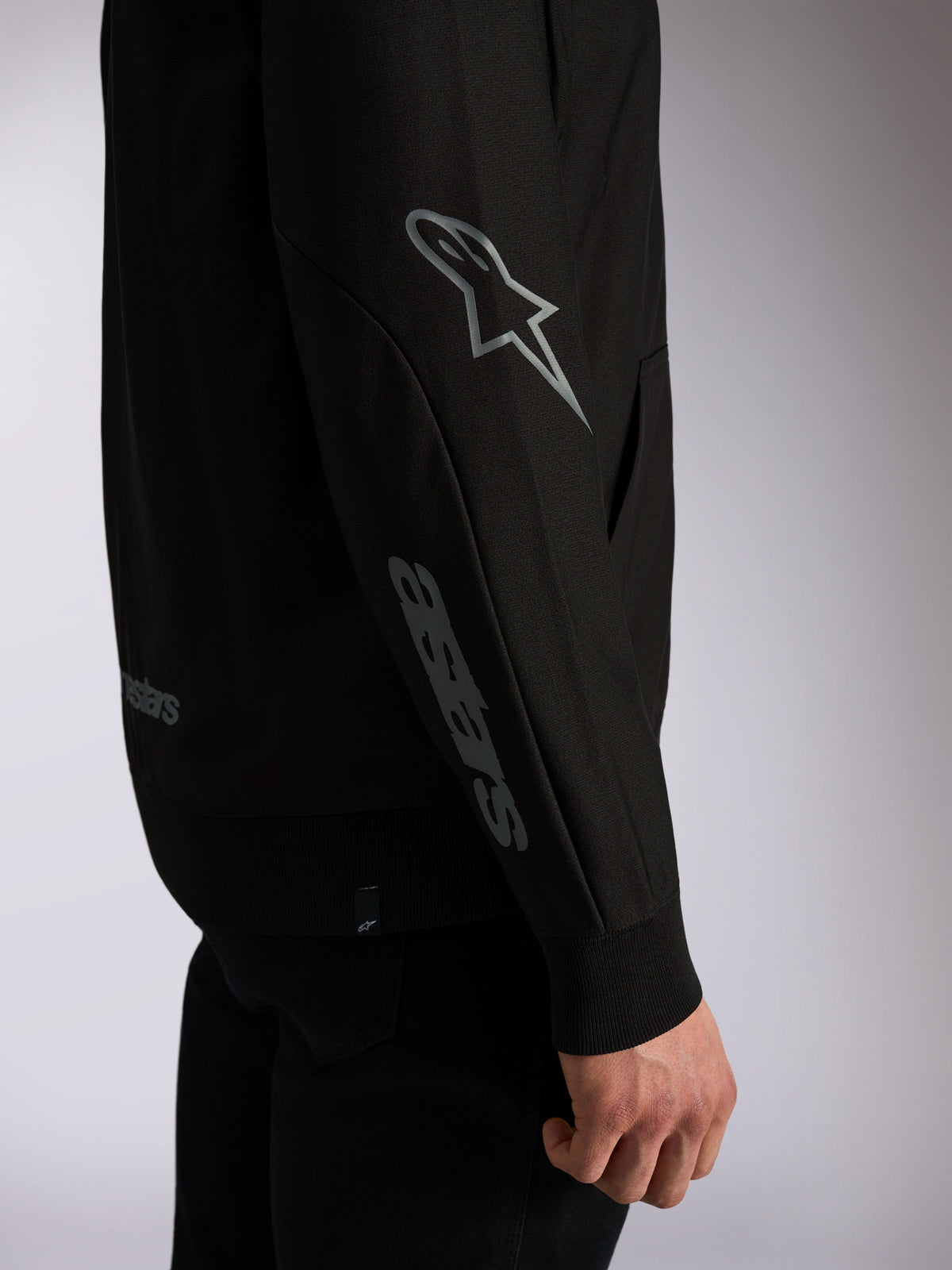 Alpinestars Sessions V3 Hoodie, schwarze Casual-Streetwear, Detailaufnahme des Ärmels mit grauem Astars-Logo am Ellbogen und Wortmarke am Unterarm, Rippbündchen, vom Motorsport inspiriertes Design