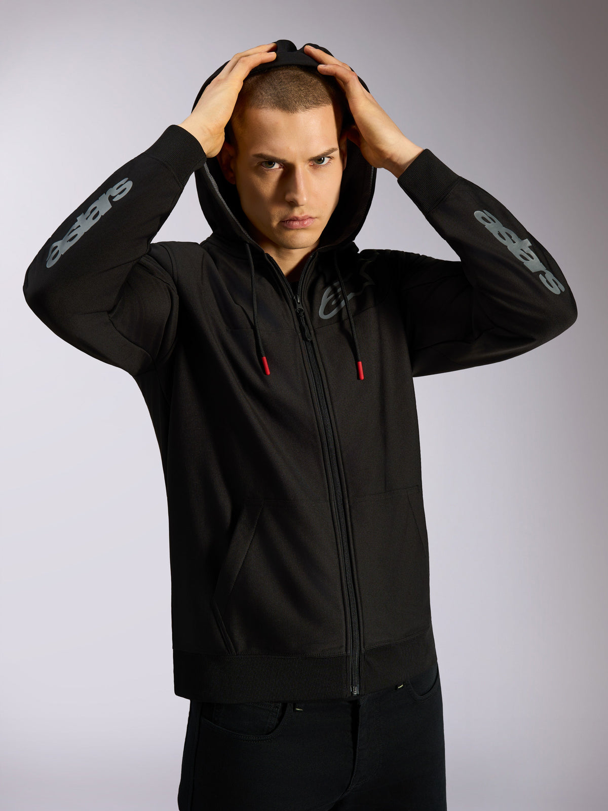 Alpinestars Sessions V3 Hoodie, schwarze Kapuzenjacke mit Reißverschluss, graues Astars-Branding auf den Unterarmen, netzgefütterte Kapuze mit Kordelzügen mit roten Enden, lässige motorsport-inspirierte Streetwear
