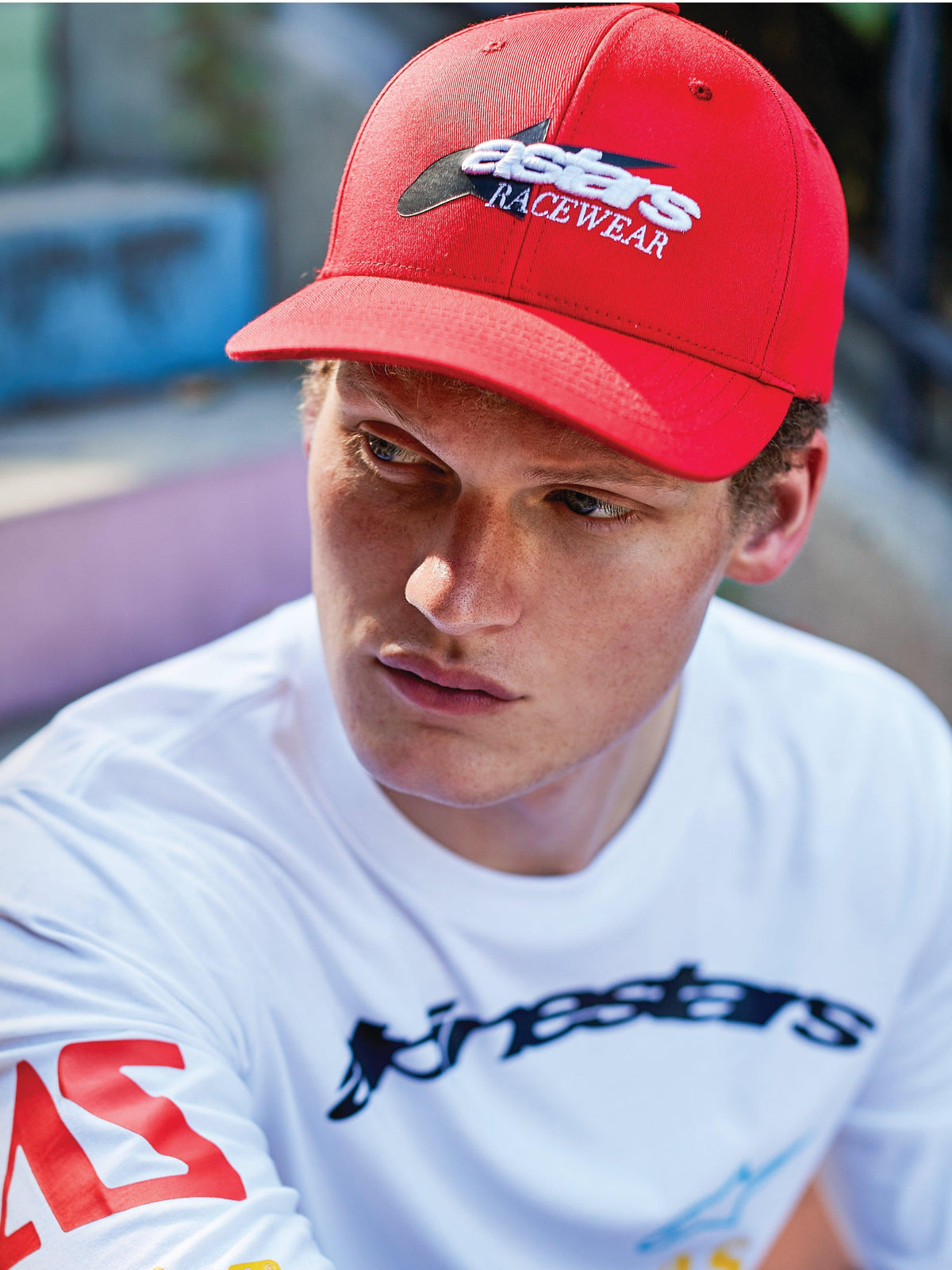 Man wearing Alpinestars Profile Hat, Red curved bill snapback, featuring white and black 'astars RACEWEAR' front embroidery, worn with a white Alpinestars long-sleeve graphic tee, casual motorsport lifestyle look