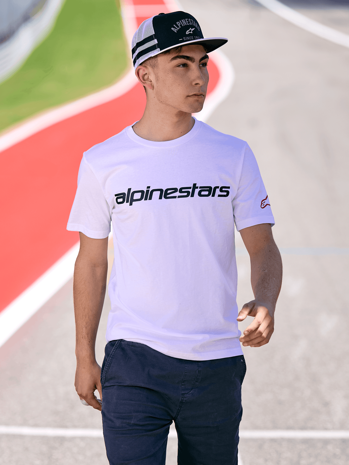 Ein Mann trägt die Alpinestars Backline Trucker Mütze in Schwarz-Weiß, kombiniert mit einem weißen Alpinestars Logo-T-Shirt und einer dunkelblauen Hose, lässige Motorsport-Streetwear vor dem Hintergrund einer Rennstrecke im Freien