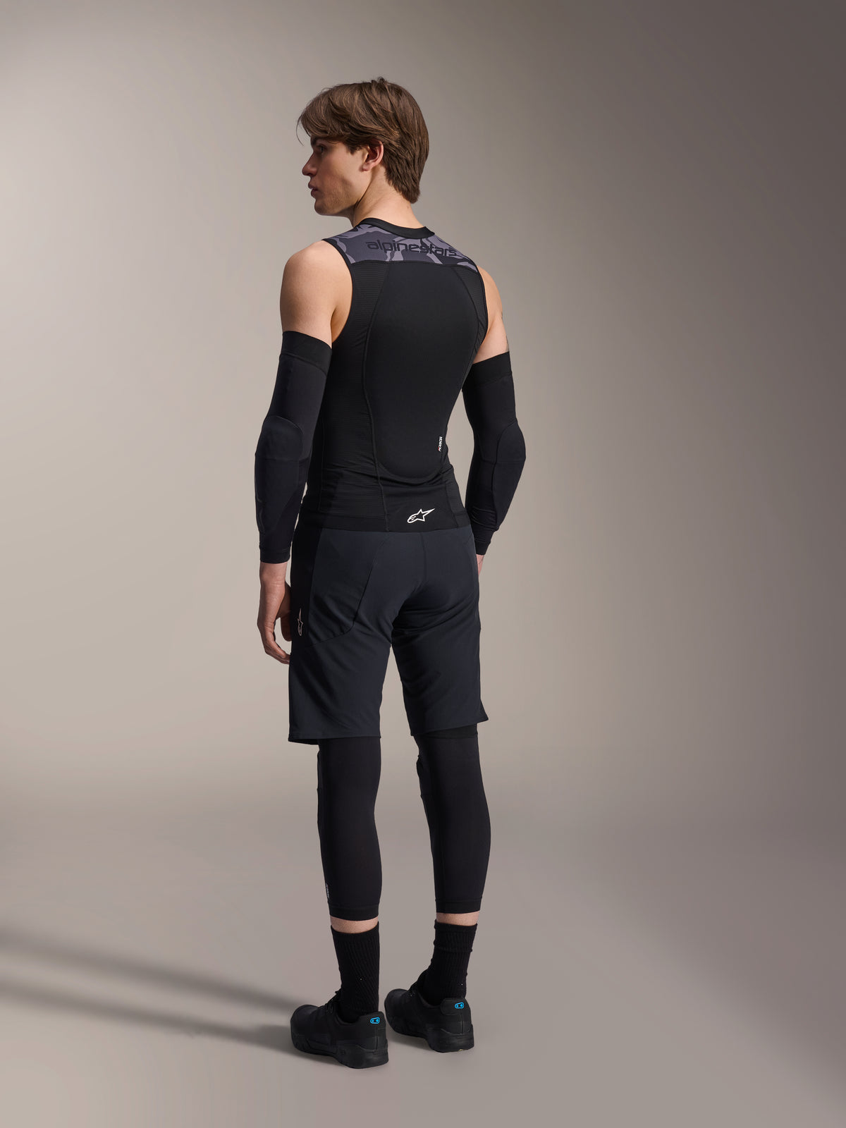 Alpinestars A-Motion Plasma Schutzweste, Black Camo, männliches Model in der Rückansicht, ärmelloses Mesh-Design mit integriertem Rückenprotektor, graues Camouflage-Muster an den Schultern, kombiniert mit schwarzen Ellbogenprotektoren und Mountainbike-Shorts, professionelle Fahrradausrüstung